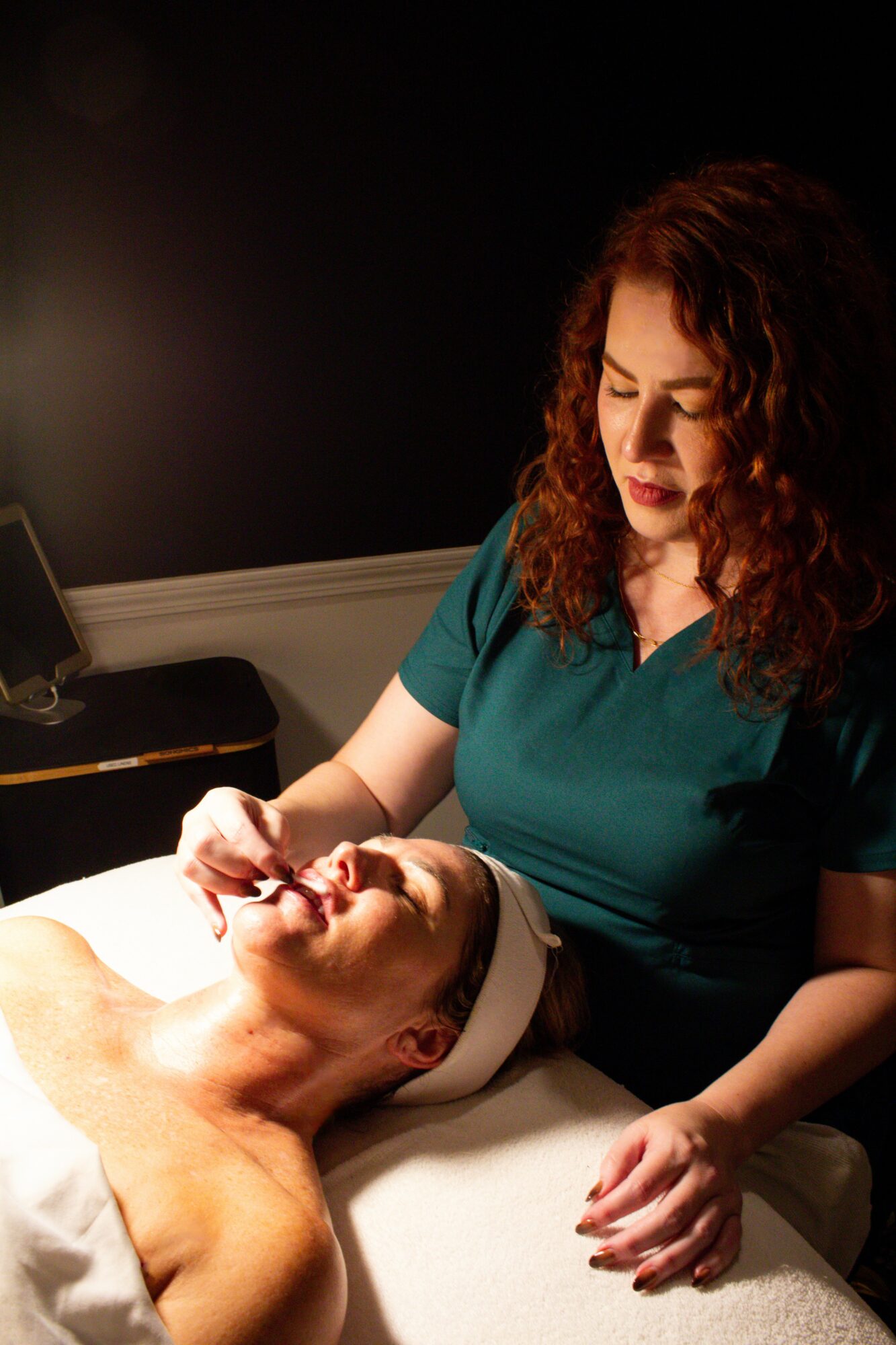 Woman with curly hair performs facial treatment on a person lying down with eyes closed.