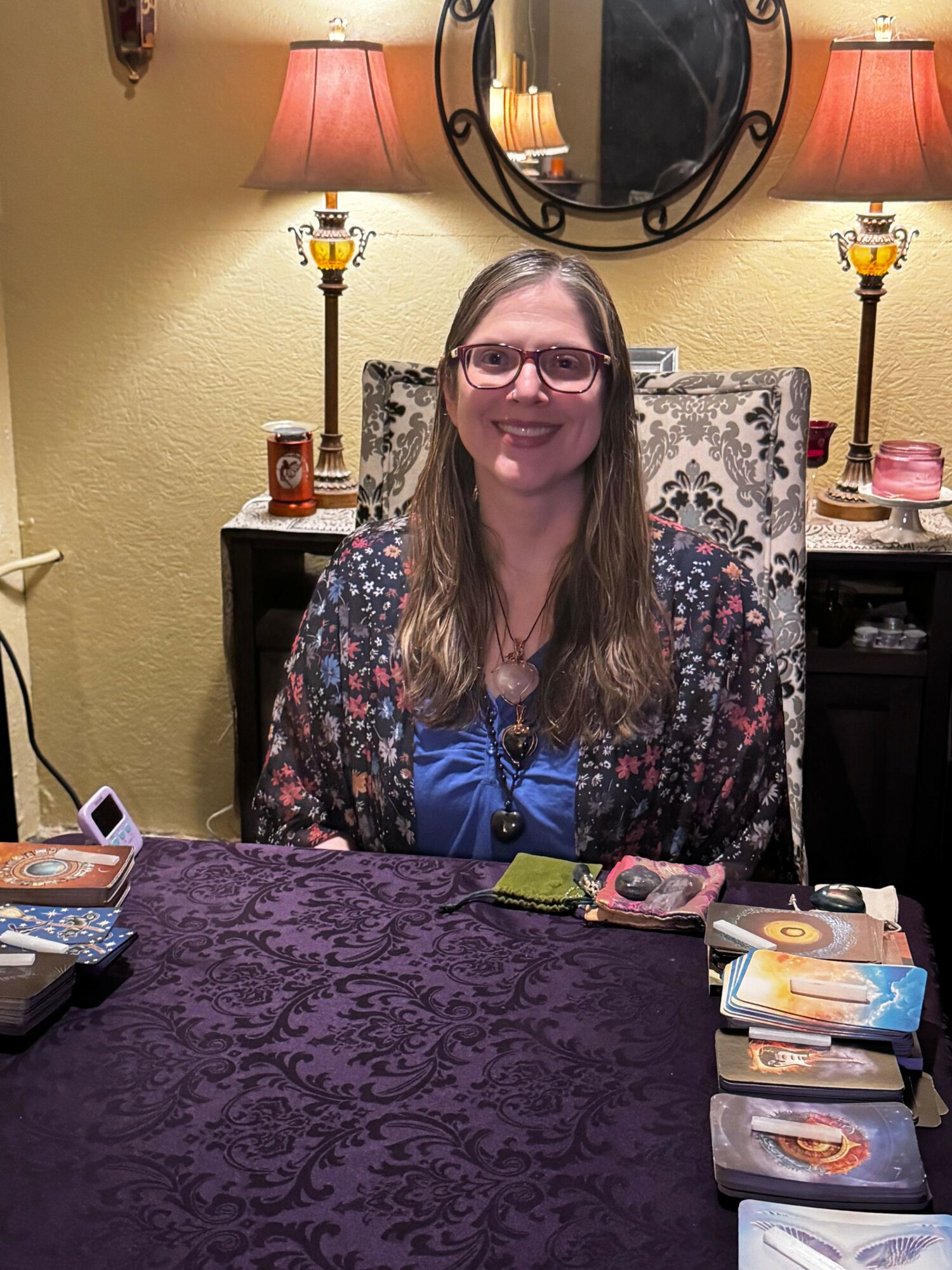 Woman with glasses and long hair sitting at a table with a patterned cloth, smiling, in a room with lamps and a mirror.