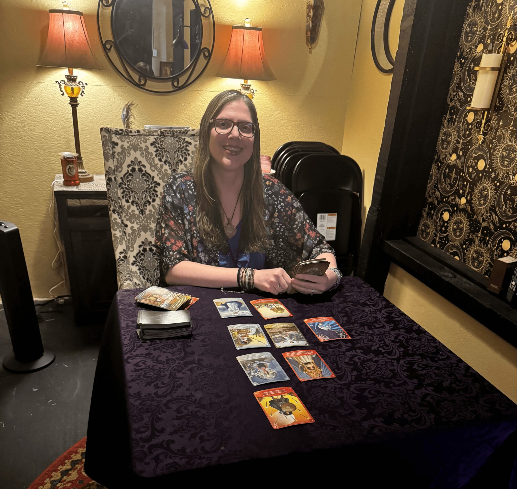 Woman sitting at a table with tarot cards, smiling, holding a card, in a warmly lit room.