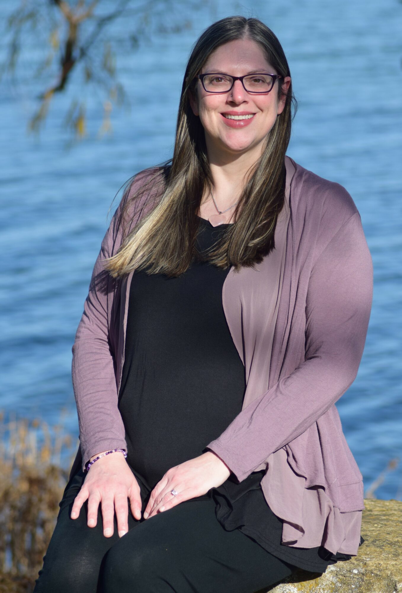 Woman with long hair and glasses smiling outdoors near water, wearing a black top and mauve cardigan.