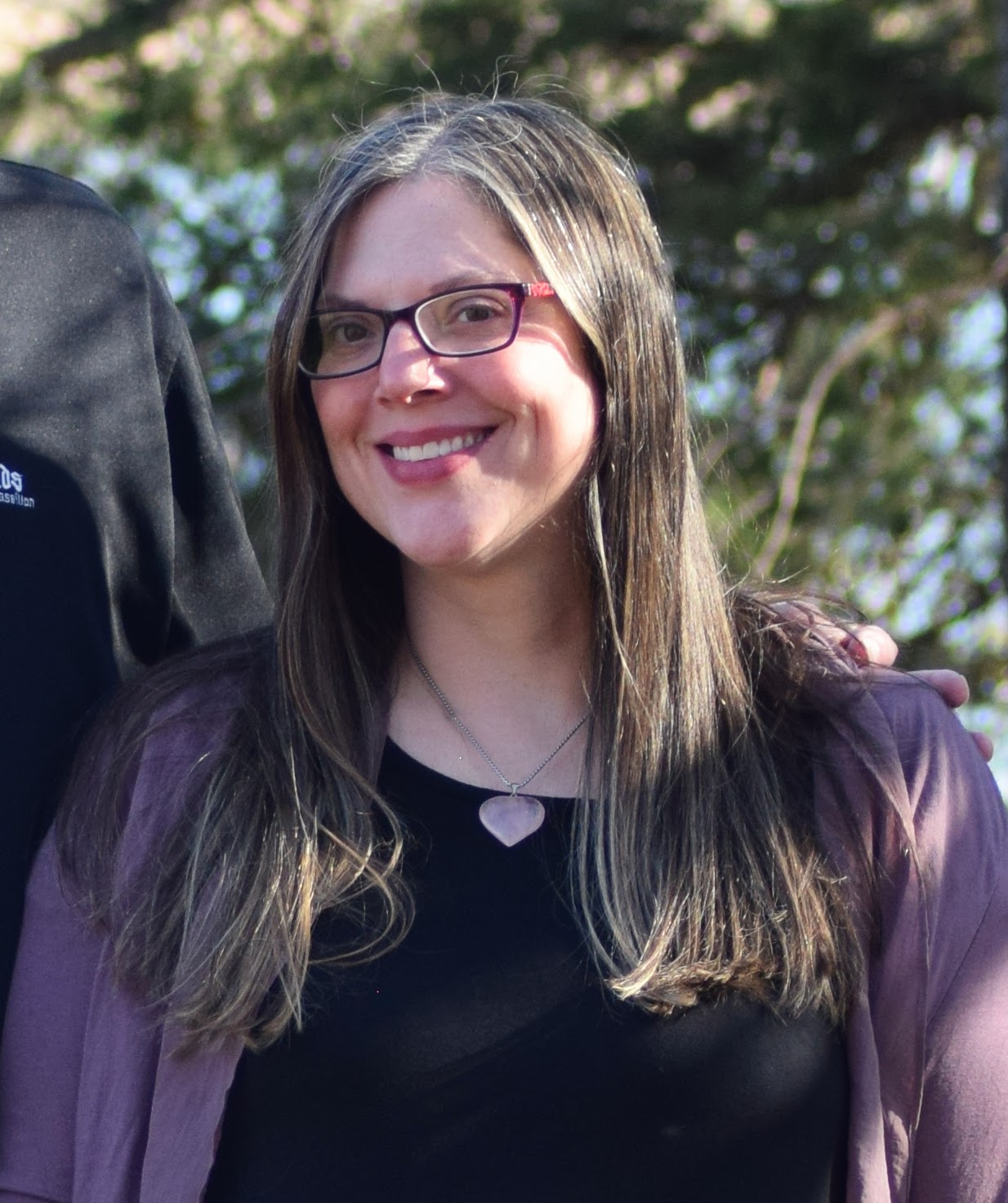 Woman with glasses smiling outdoors, wearing a purple jacket and black shirt, with trees in background.