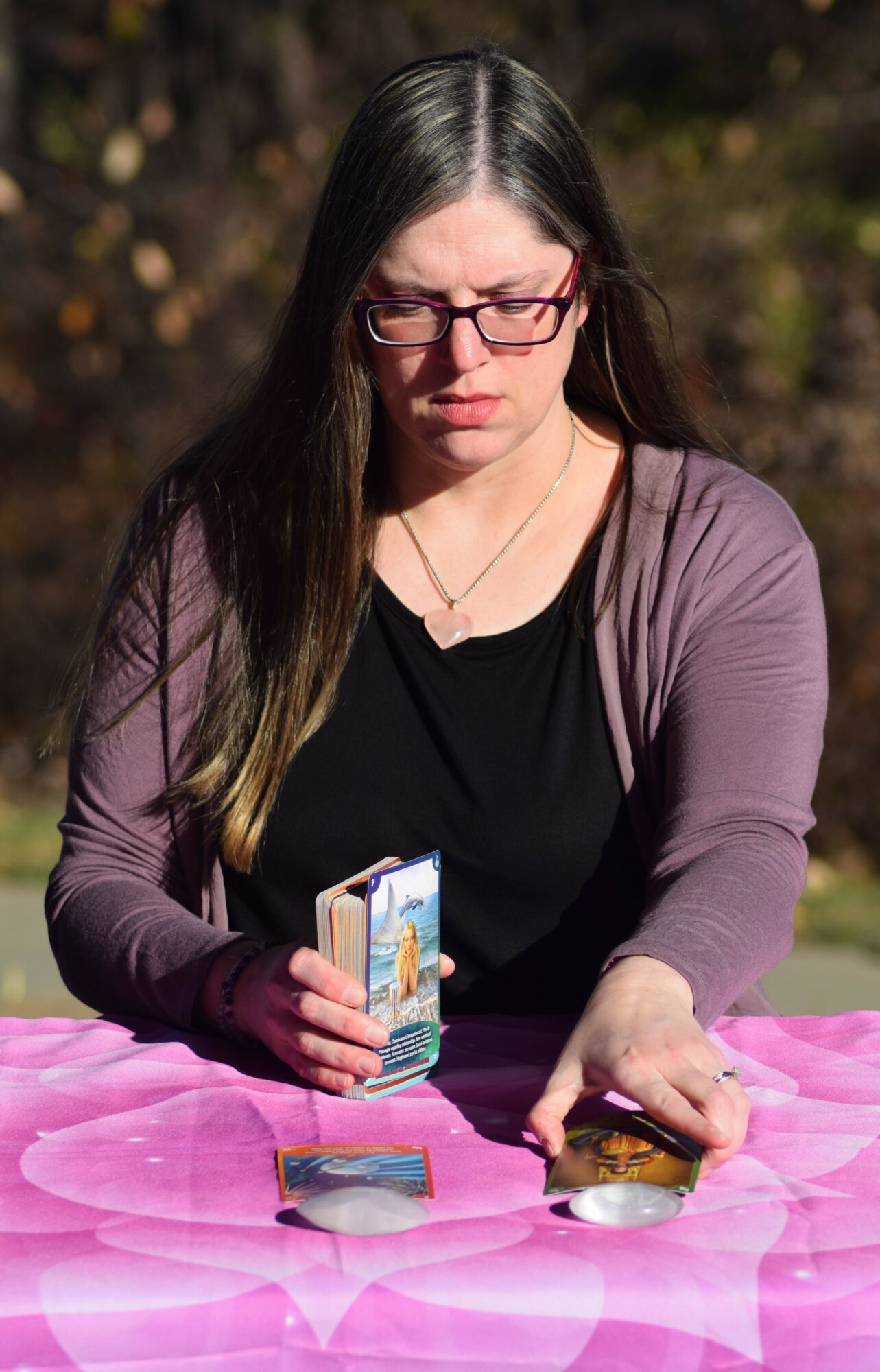 Woman with long dark hair and glasses, wearing a black shirt and purple cardigan, looking at tarot cards on pink cloth.