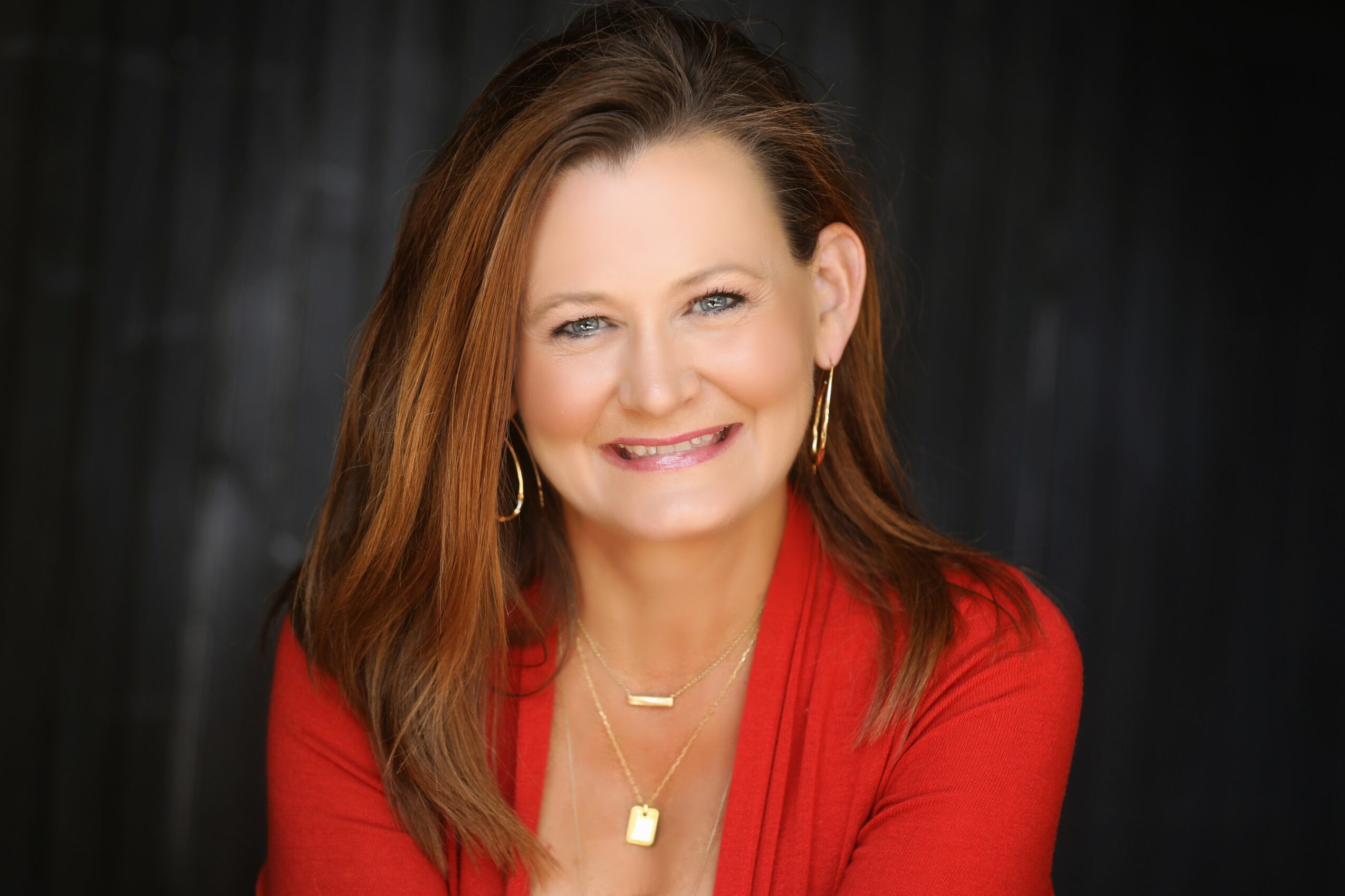 Smiling woman with shoulder-length brown hair wearing a red top and gold jewelry.