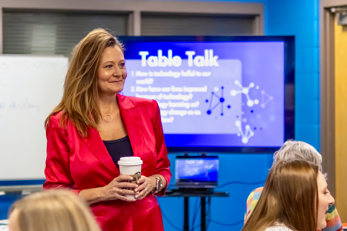 Woman in red blazer holding coffee cup presents in front of a screen with a presentation slide titled 'Table Talk'.