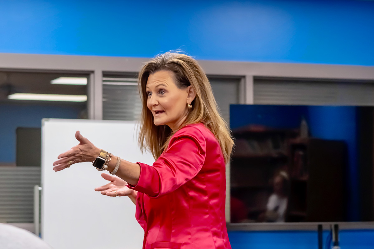Woman in pink jacket gesturing with hands in a modern indoor setting.