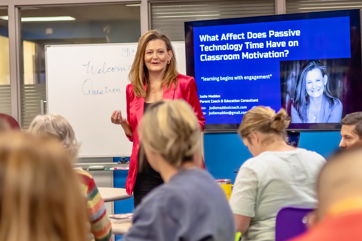 Woman in red blazer standing in front of classroom with students, presentation slide on screen behind her.