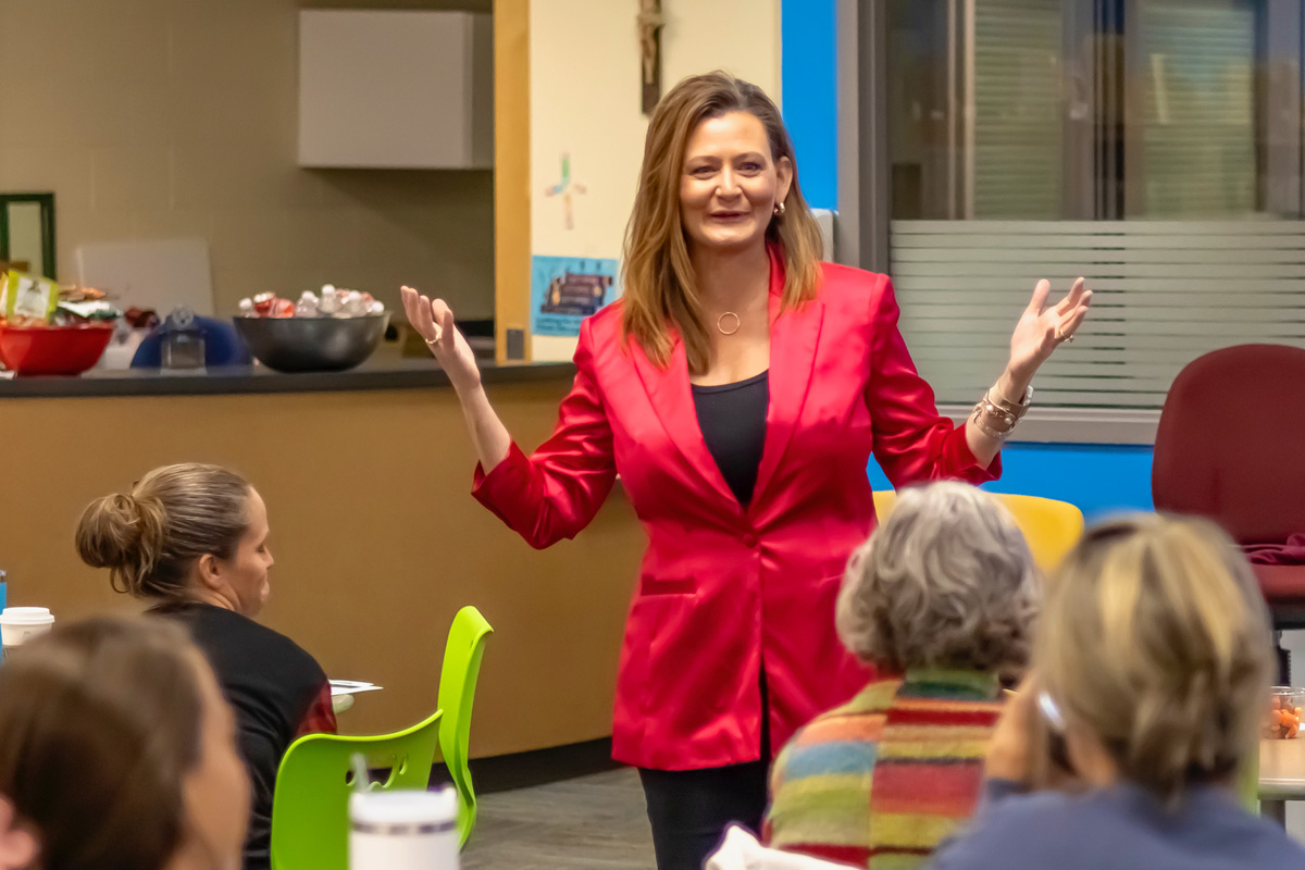 Woman in red blazer speaking to a group of people in a room with colorful chairs and a counter.