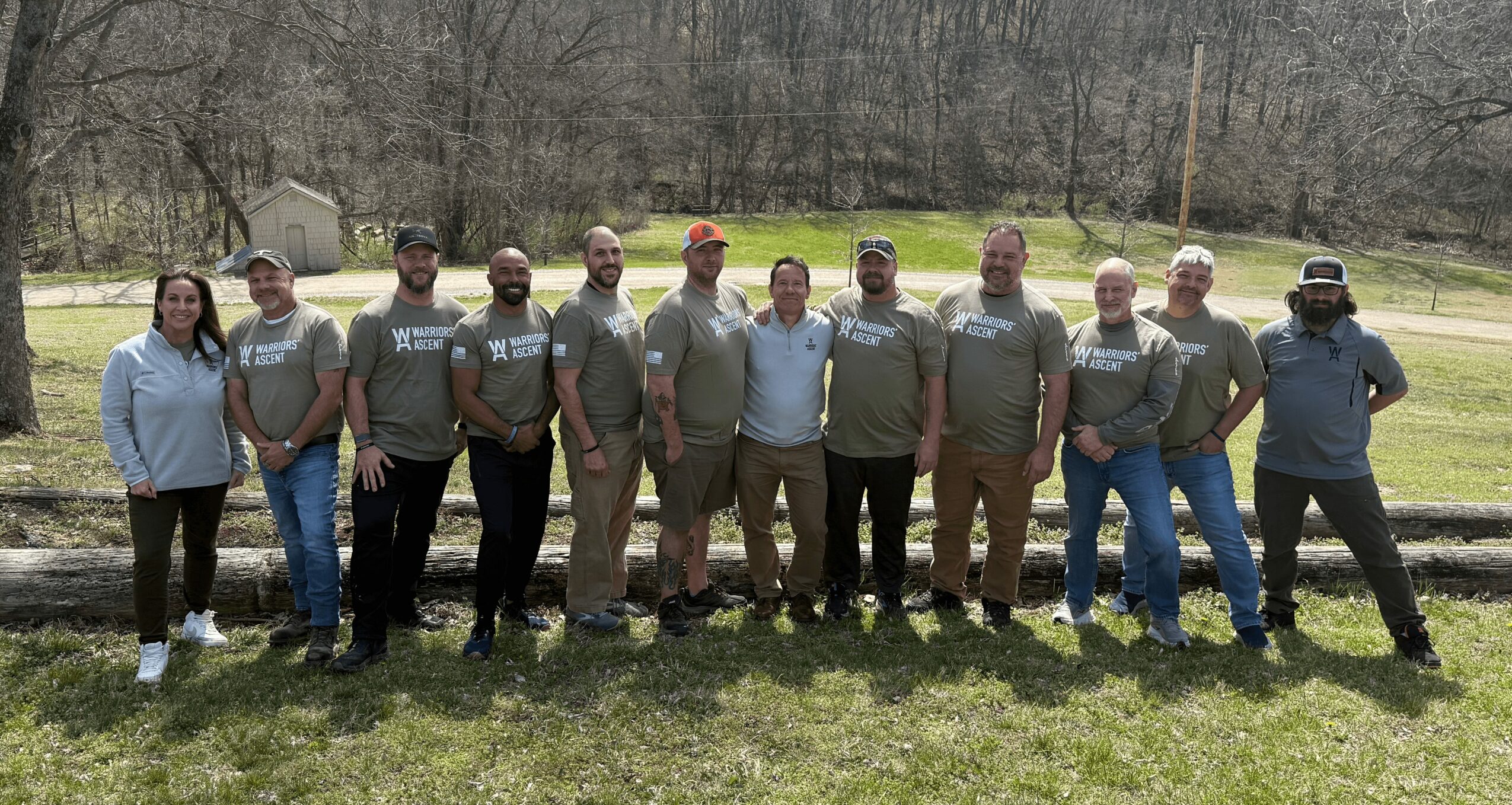 Group of eleven people standing outdoors on grass, some wearing matching shirts, in a park with trees and a road behind.