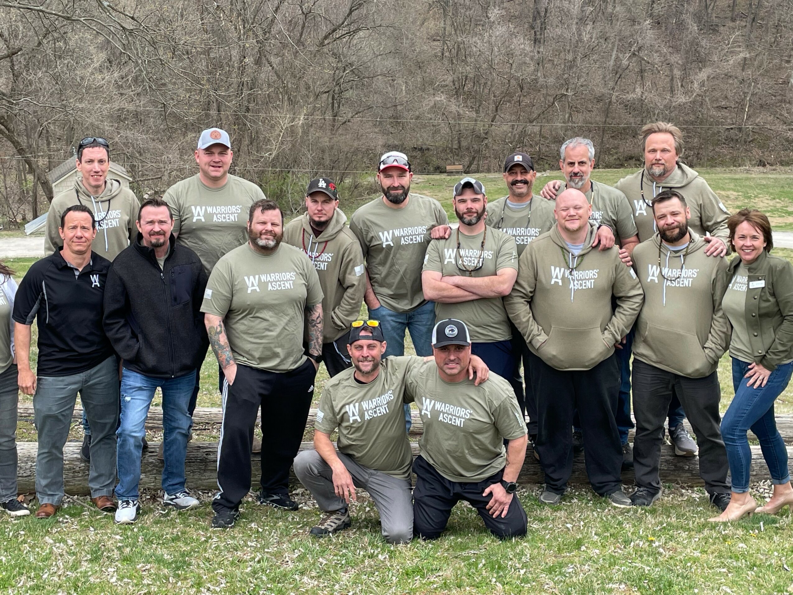 Group of 16 people outdoors on grass, some wearing matching gray shirts, in a wooded area.