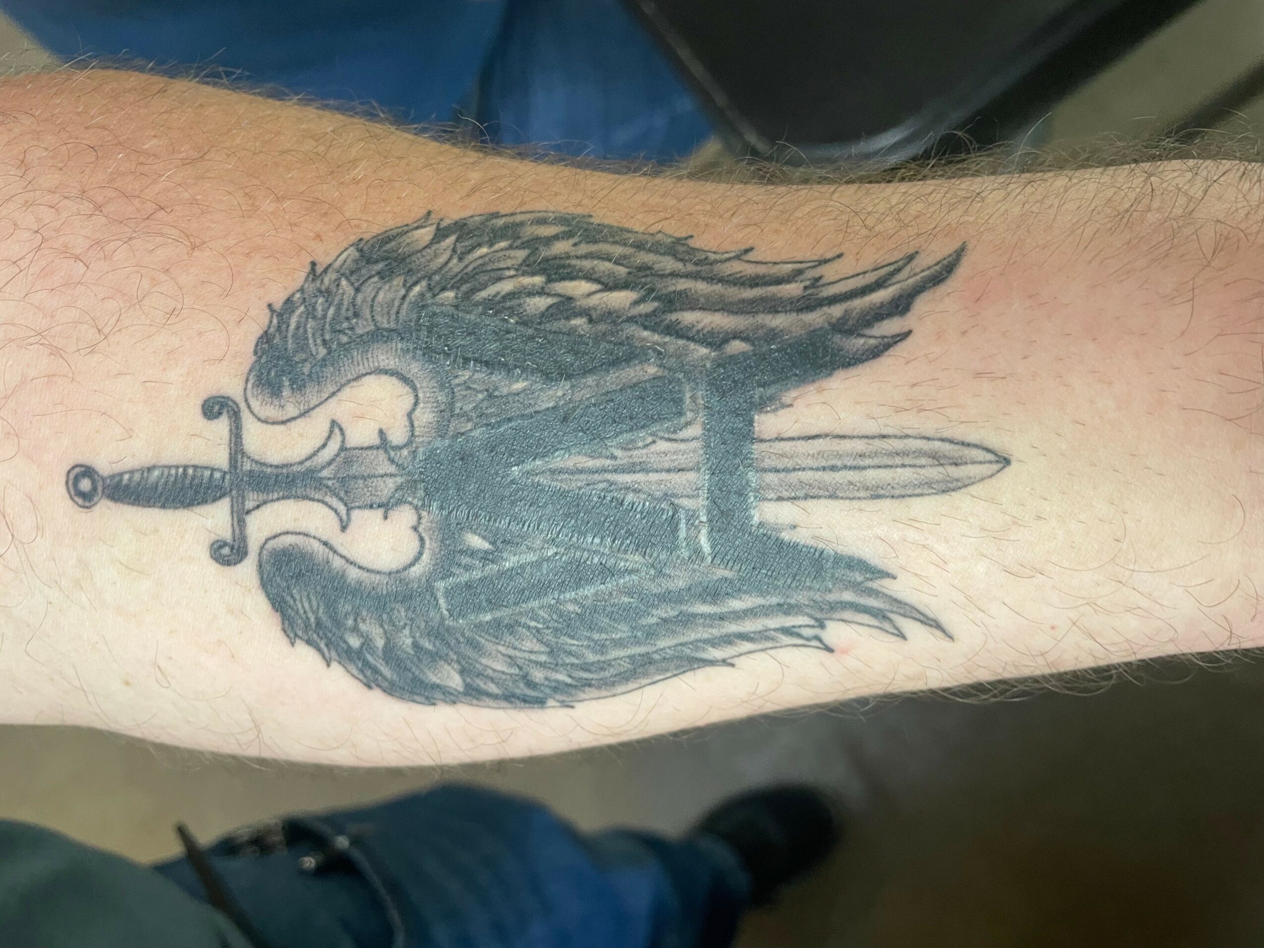 Black and gray tattoo of a sword with wings on a person's forearm.