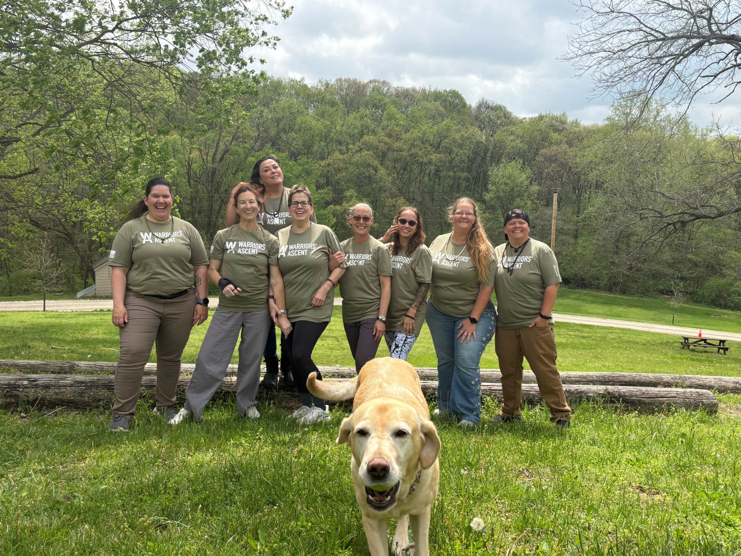 Group of seven people standing outdoors with a dog in front, trees and grass in background.