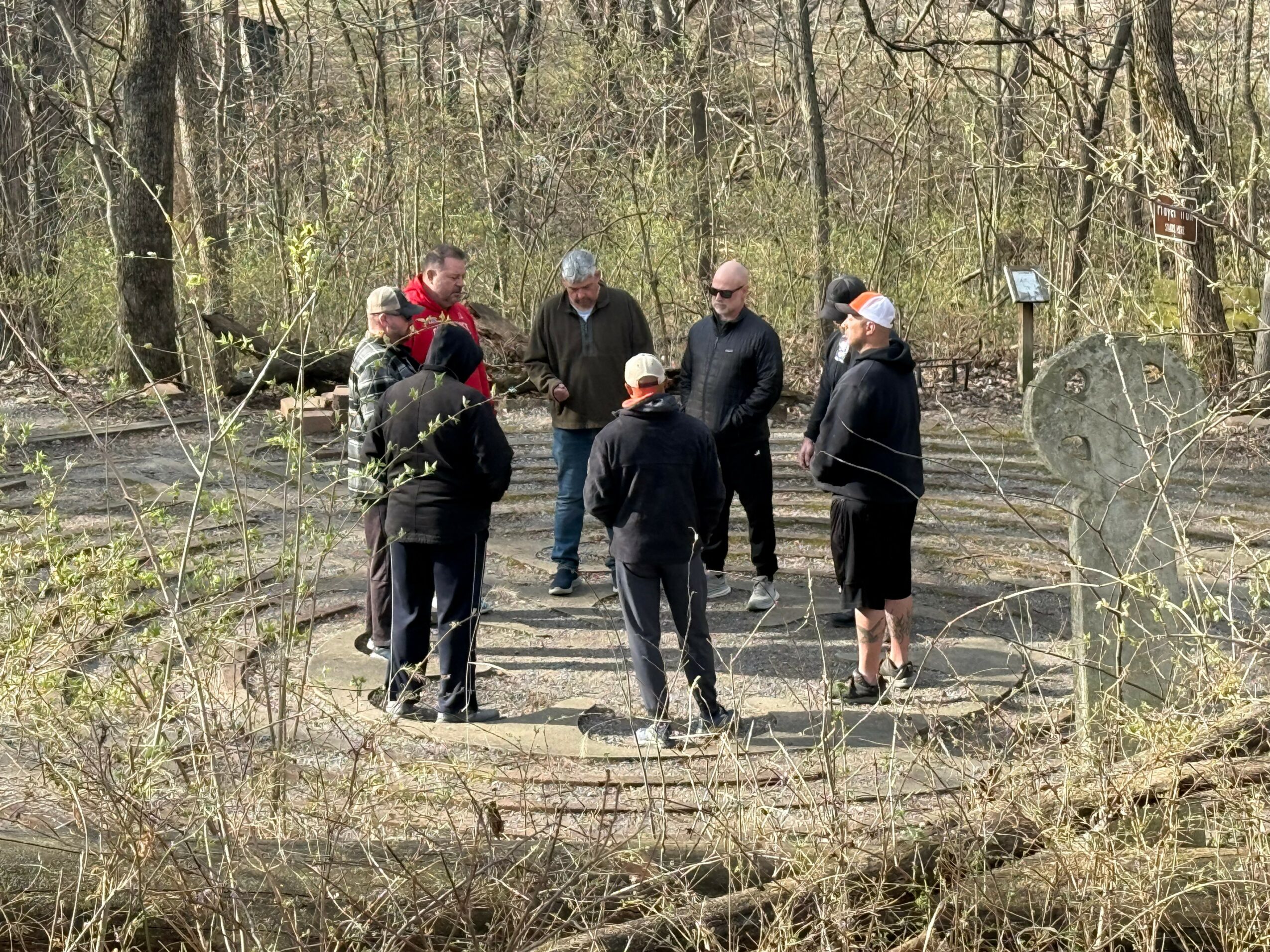 Group of seven people standing and talking outdoors in a wooded area with trees and a path.