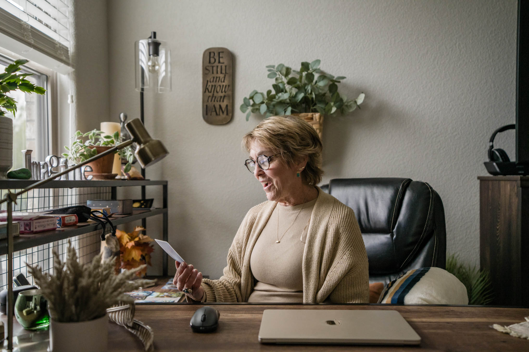 Woman with glasses sitting at desk, reading a paper, in a room with plants and office supplies.
