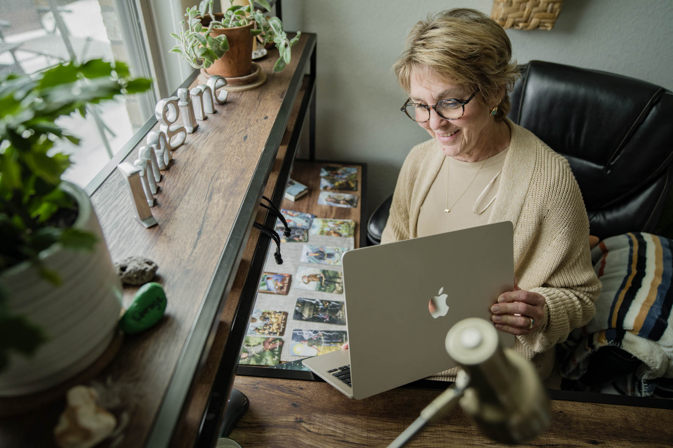 Older woman with glasses using a laptop in a cozy room with plants and photos on the table.