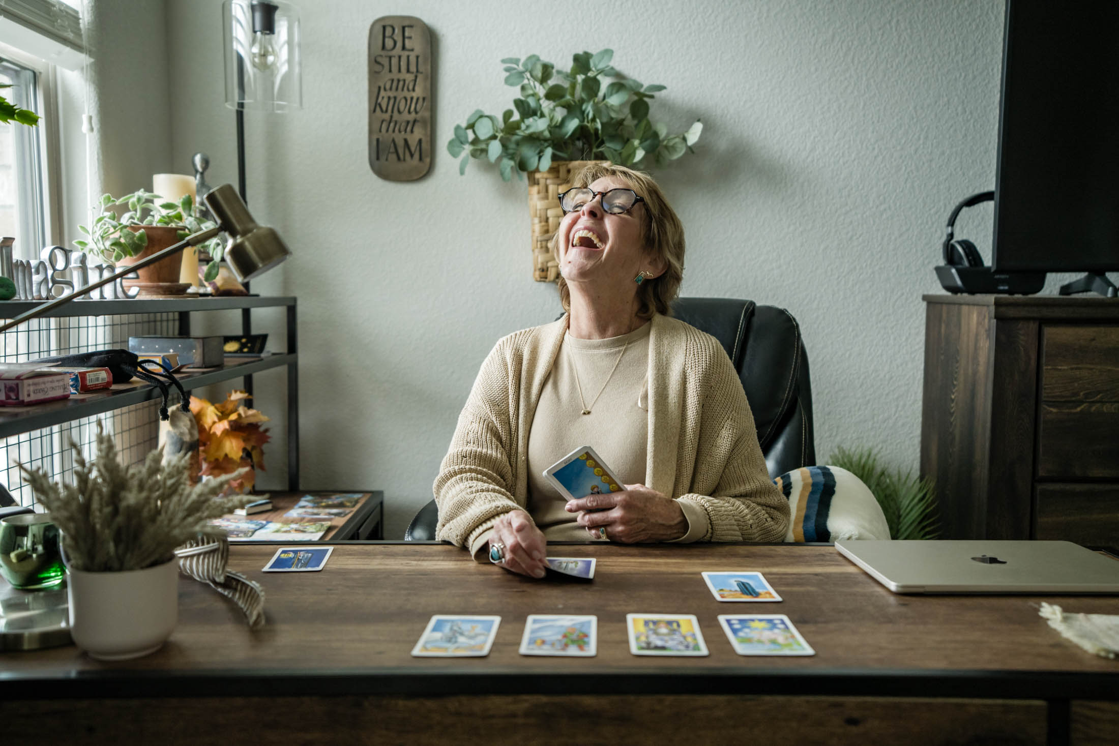 Woman sitting at desk laughing, holding a phone, with cards spread on desk, plants, and wall decorations in background.