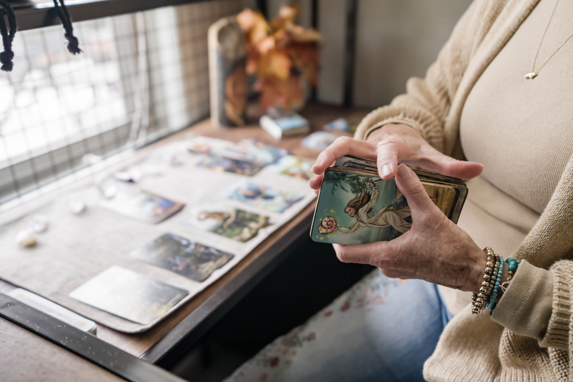 Person holding a tarot card deck with a table of cards and a window in the background.