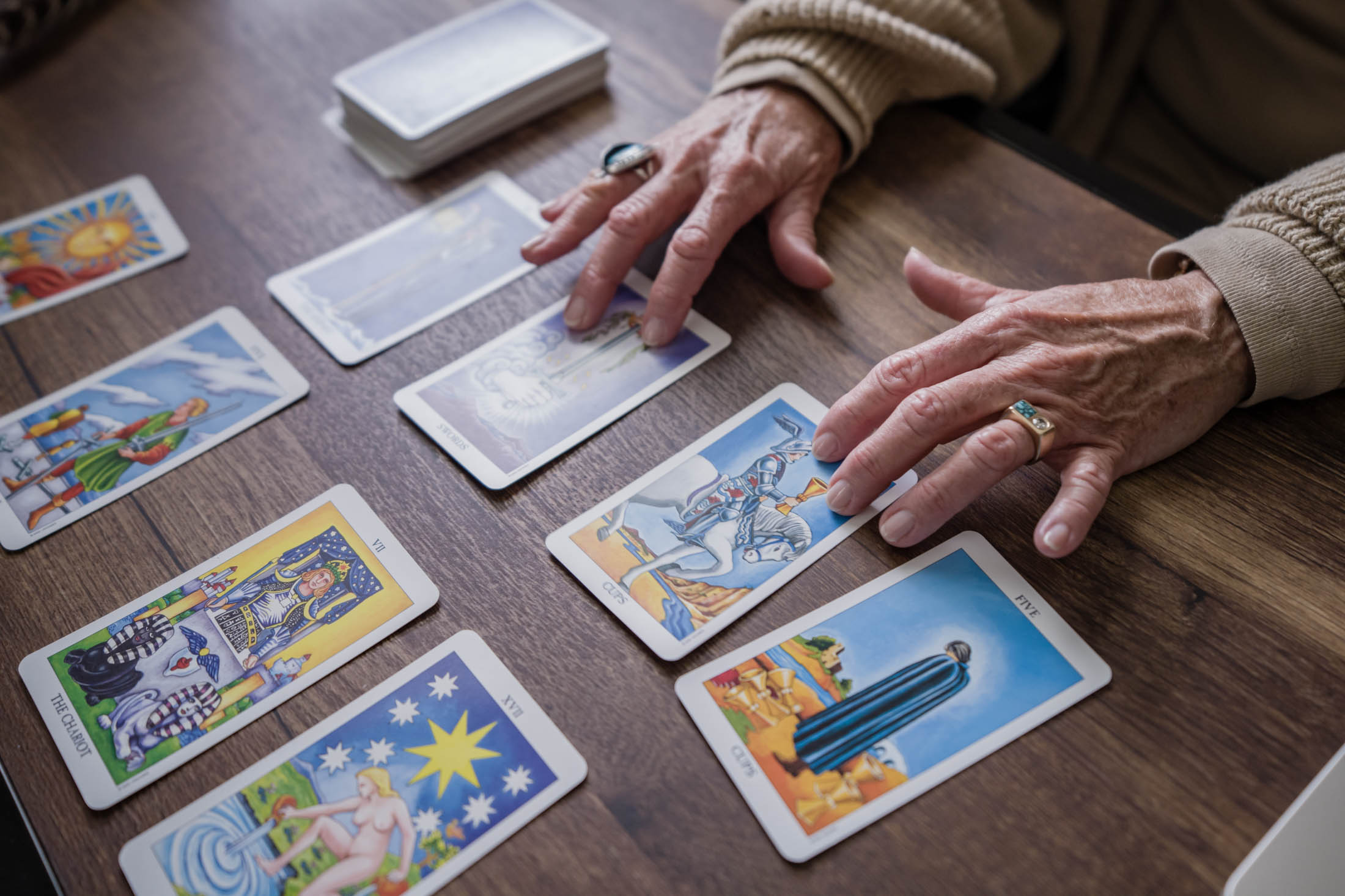 Person's hands arranging tarot cards on a wooden table, with additional cards nearby, wearing a ring and a beige sweater.