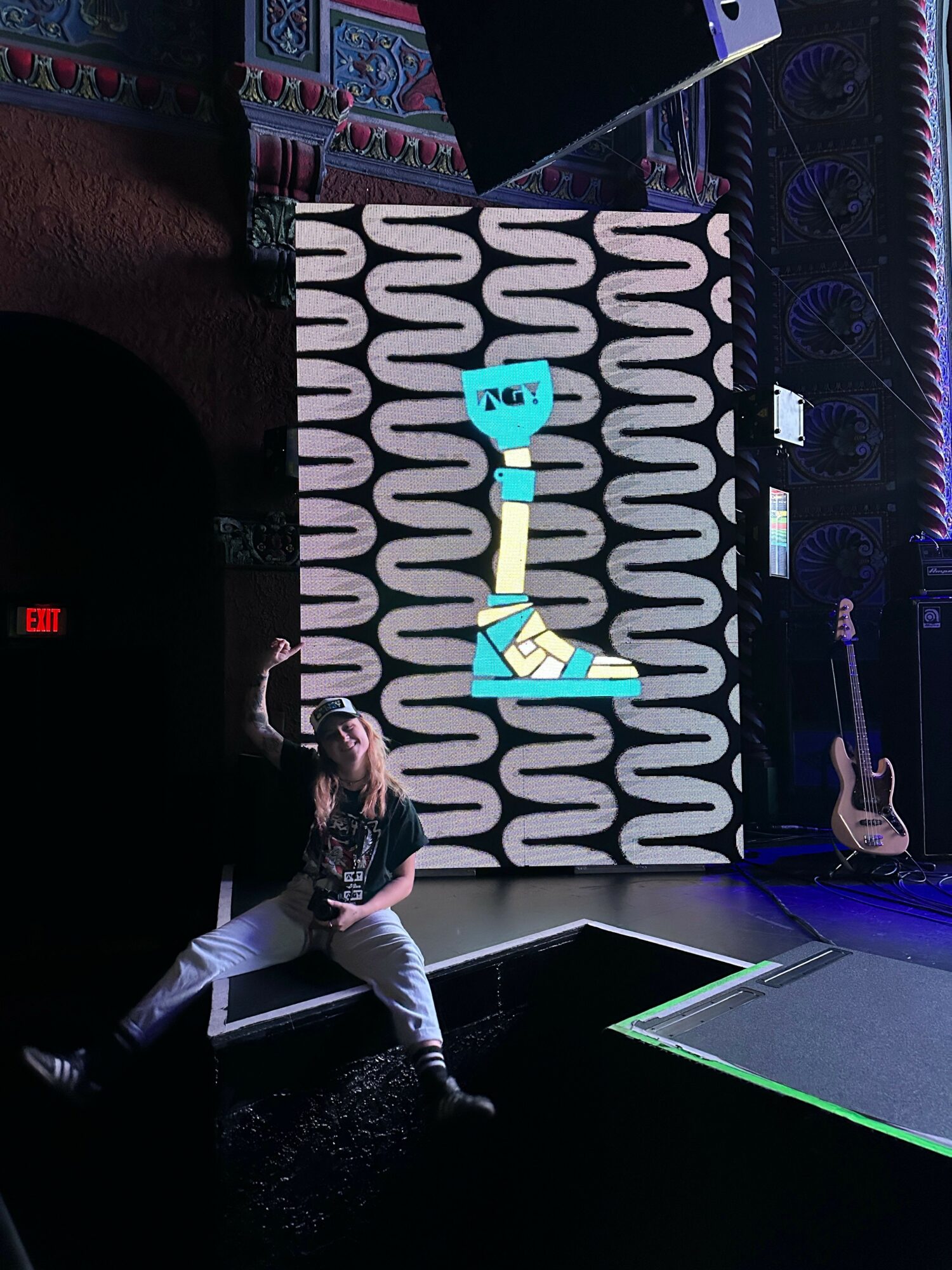 Person performing on stage with a large patterned backdrop, guitar, and stage lighting.