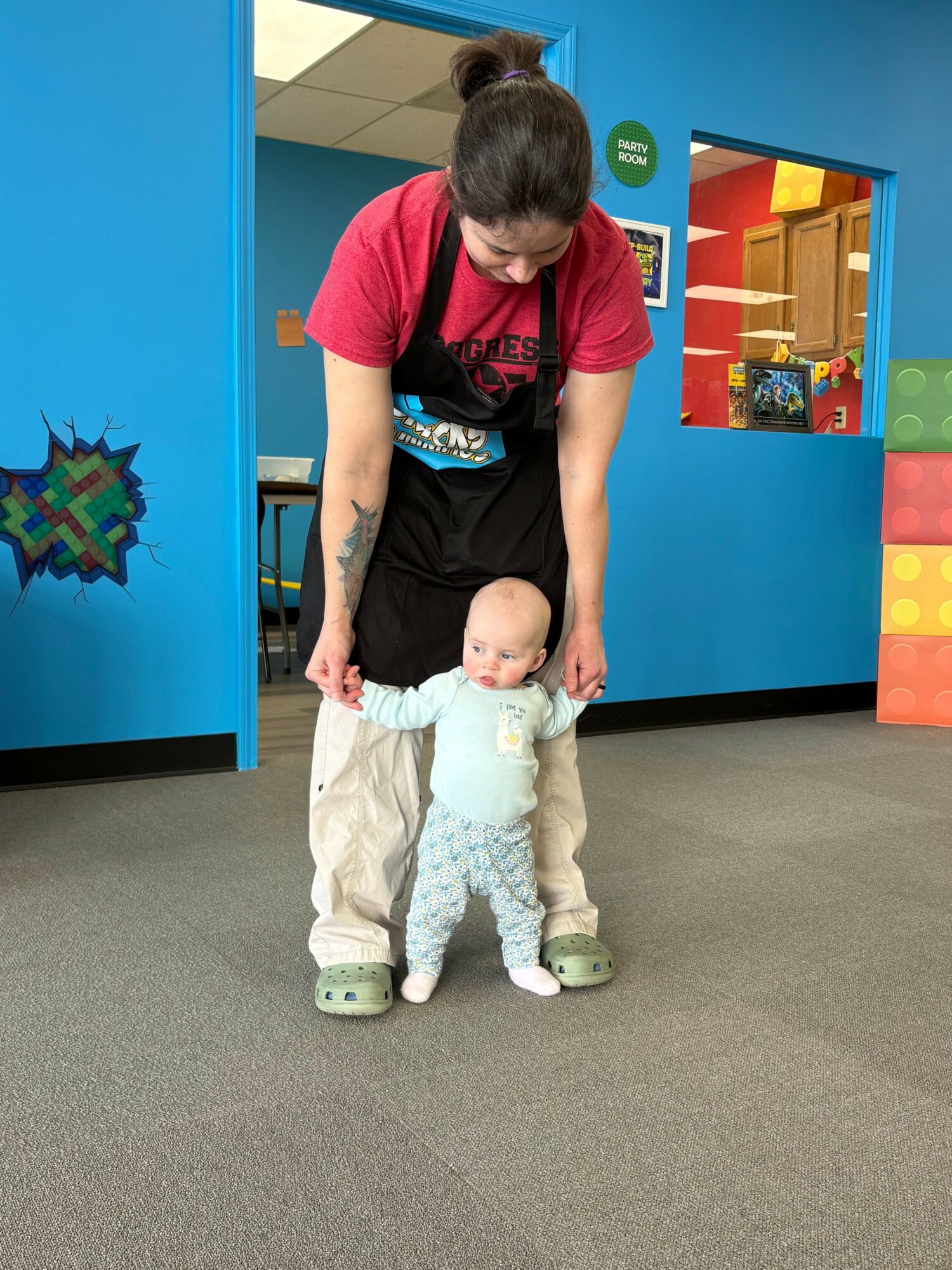 Adult holding a baby's hands in a colorful indoor play area, blue walls, and foam blocks in background.