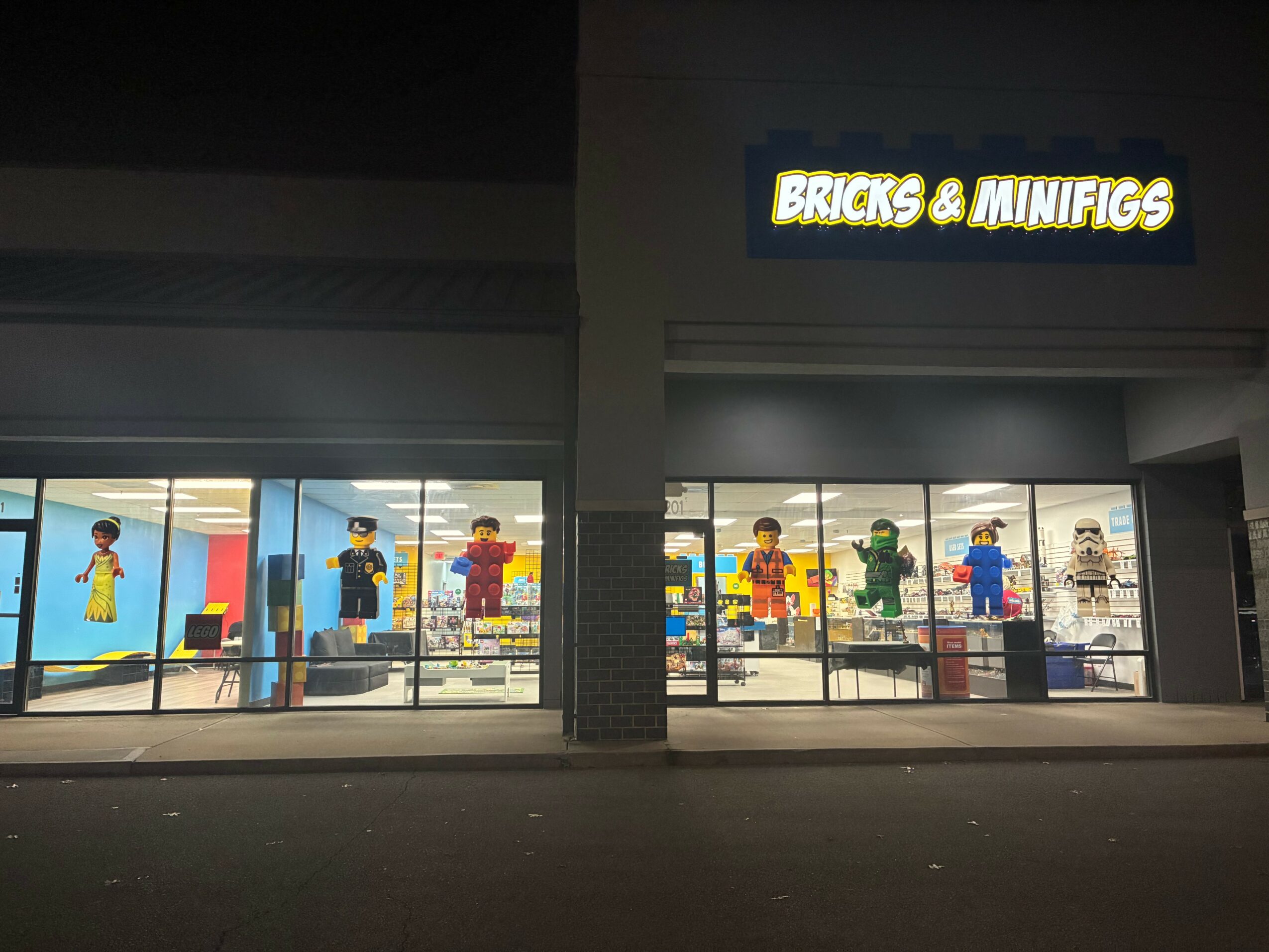 Store window display with LEGO minifigures inside, illuminated sign reads 'BRICKS & MINIFIGS'.