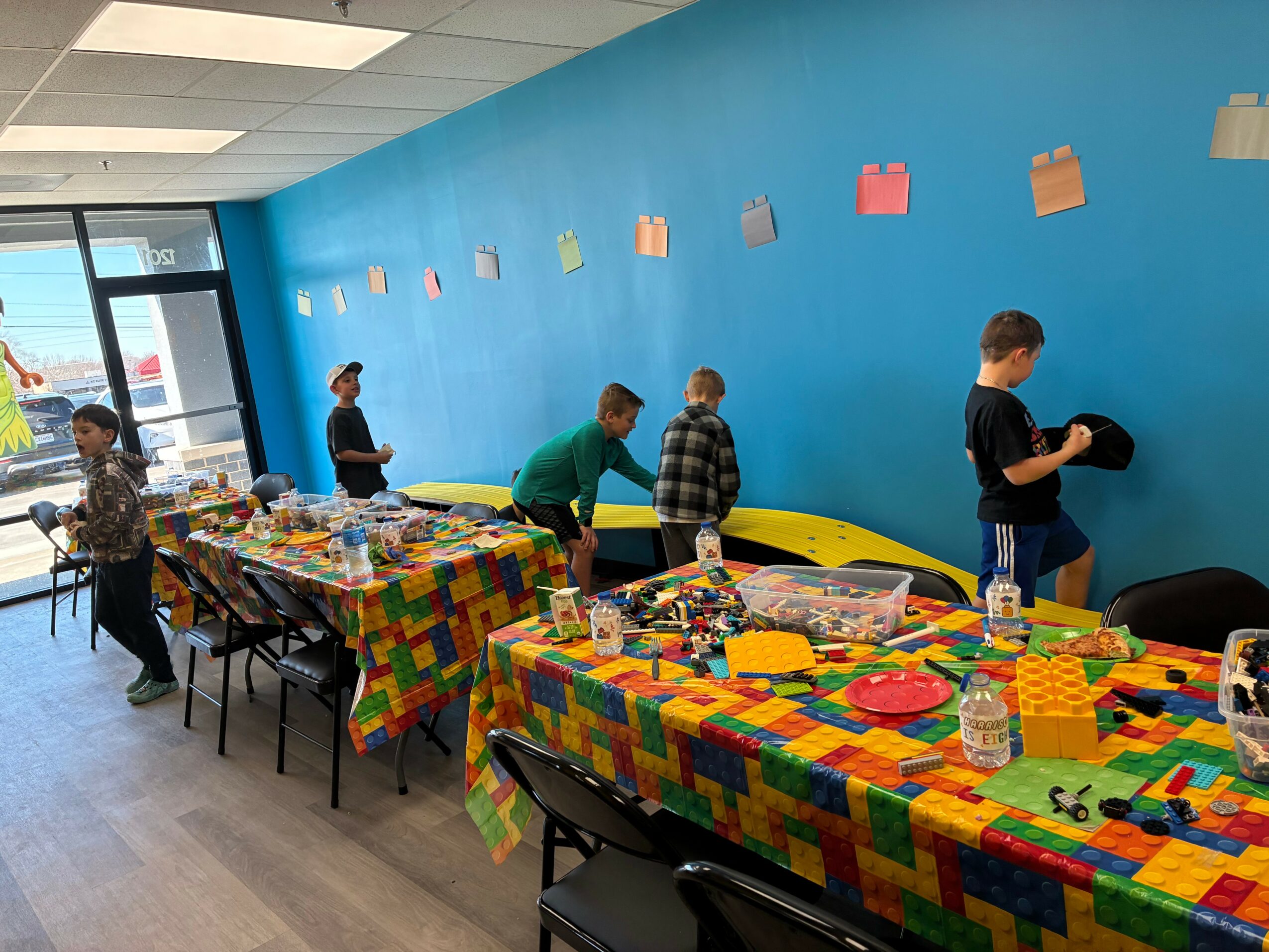 Children and adults in a room with a blue wall, colorful tables, and chairs, engaging in activities.