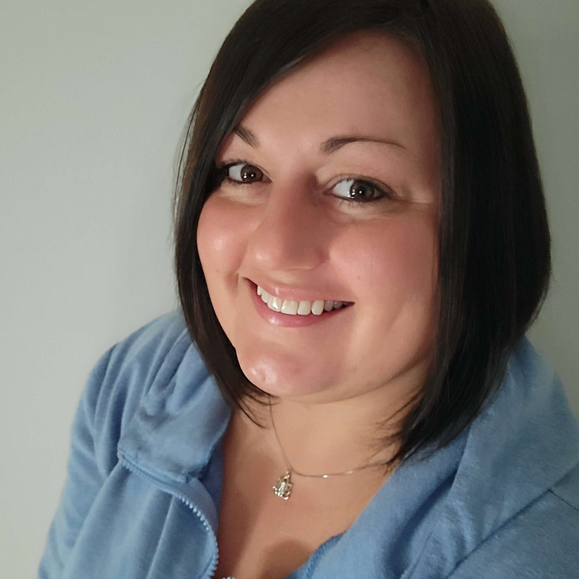 Smiling woman with shoulder-length dark hair, wearing a blue top and a necklace, against a plain background.