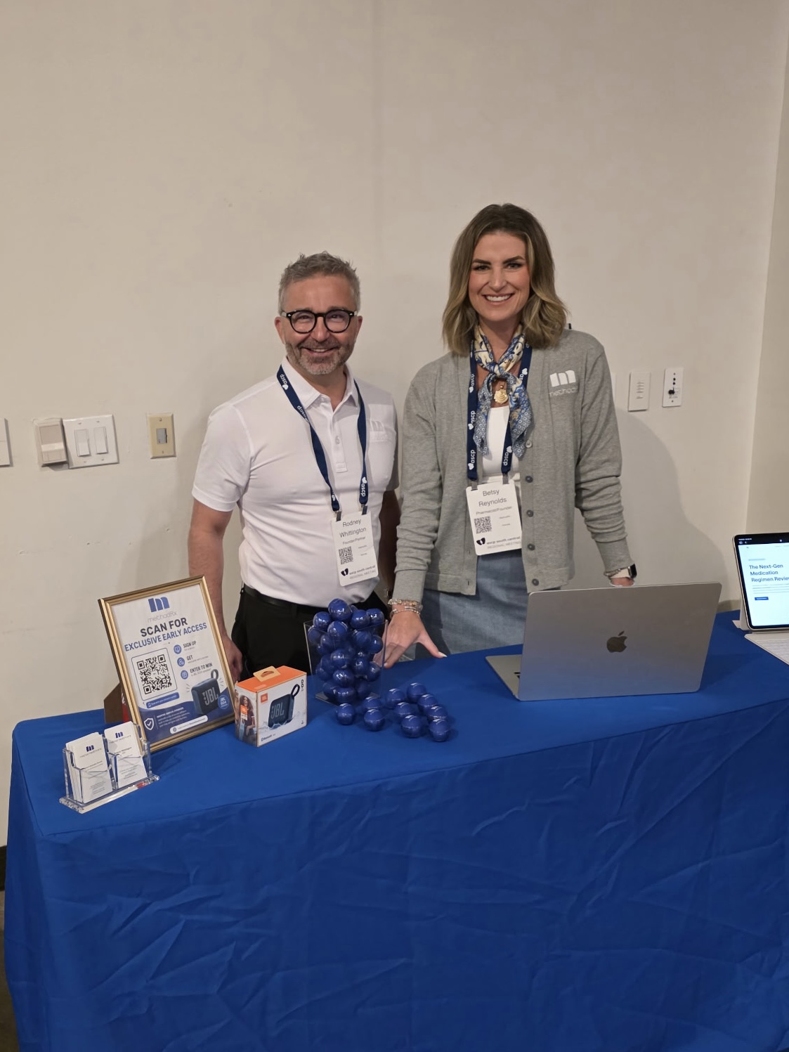 Two people standing behind a table with promotional items, smiling, in a plain room.