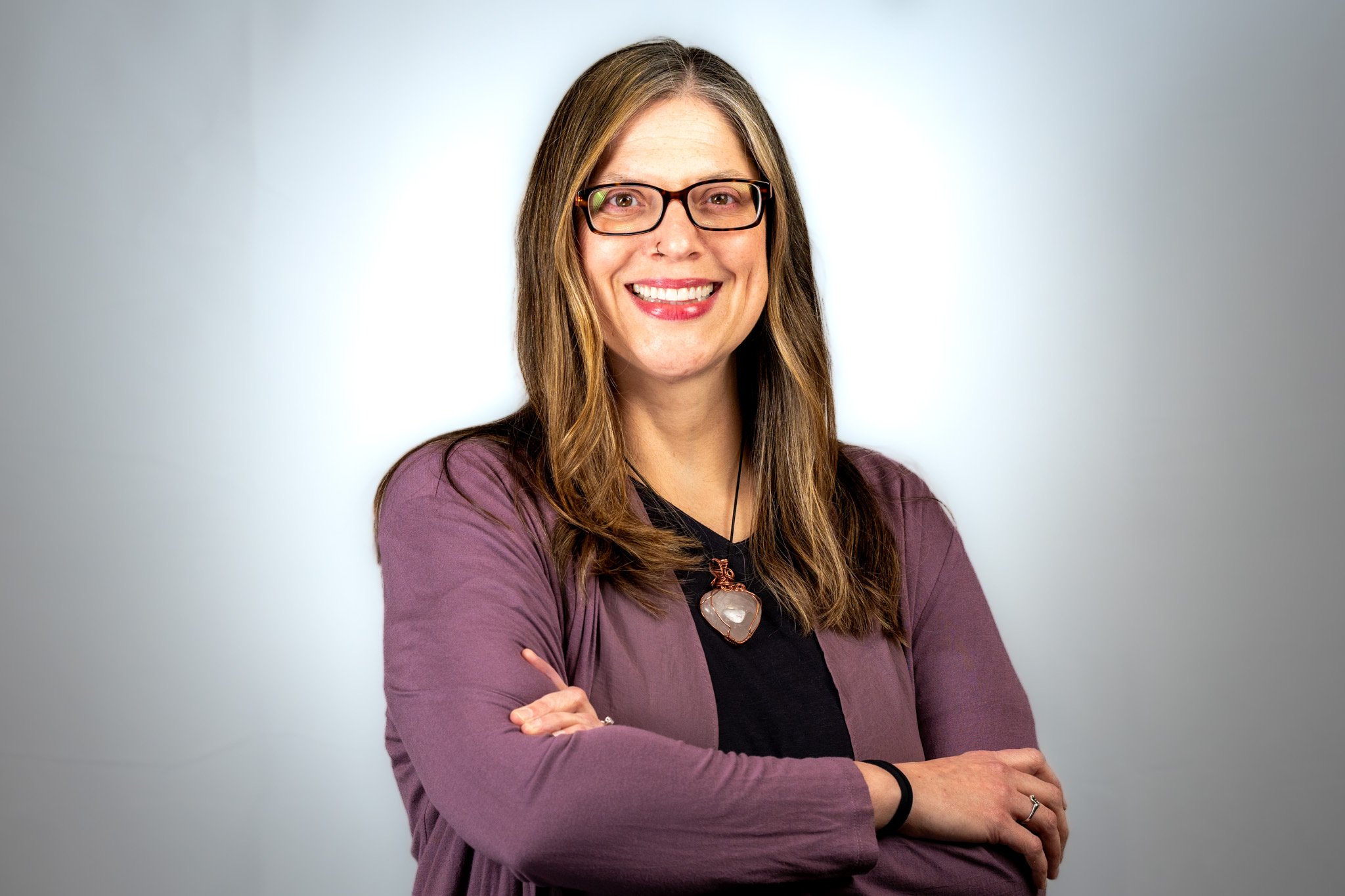 Woman with glasses smiling, arms crossed, wearing a purple blazer and black top, standing against a light background.