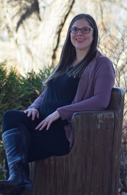 Woman with glasses sitting on a wooden bench outdoors, smiling, wearing a purple cardigan and black outfit.