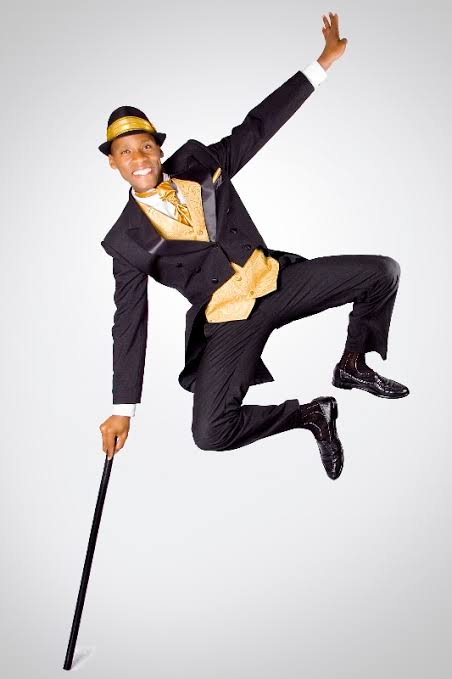 Person in a suit and hat jumping with one arm raised, holding a cane, against a plain background.