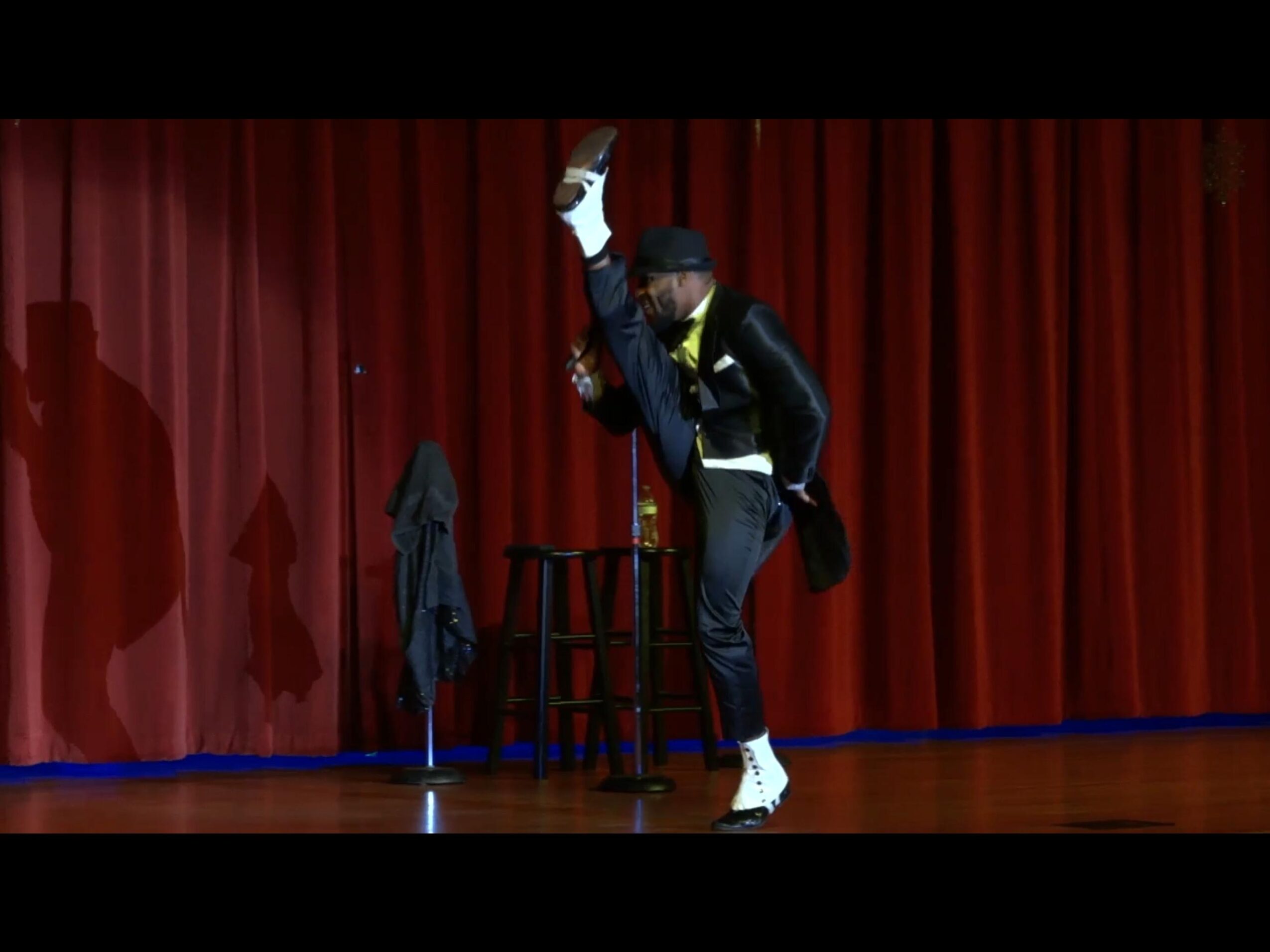 Performer in black outfit and white shoes doing a high kick on stage with red curtains behind.