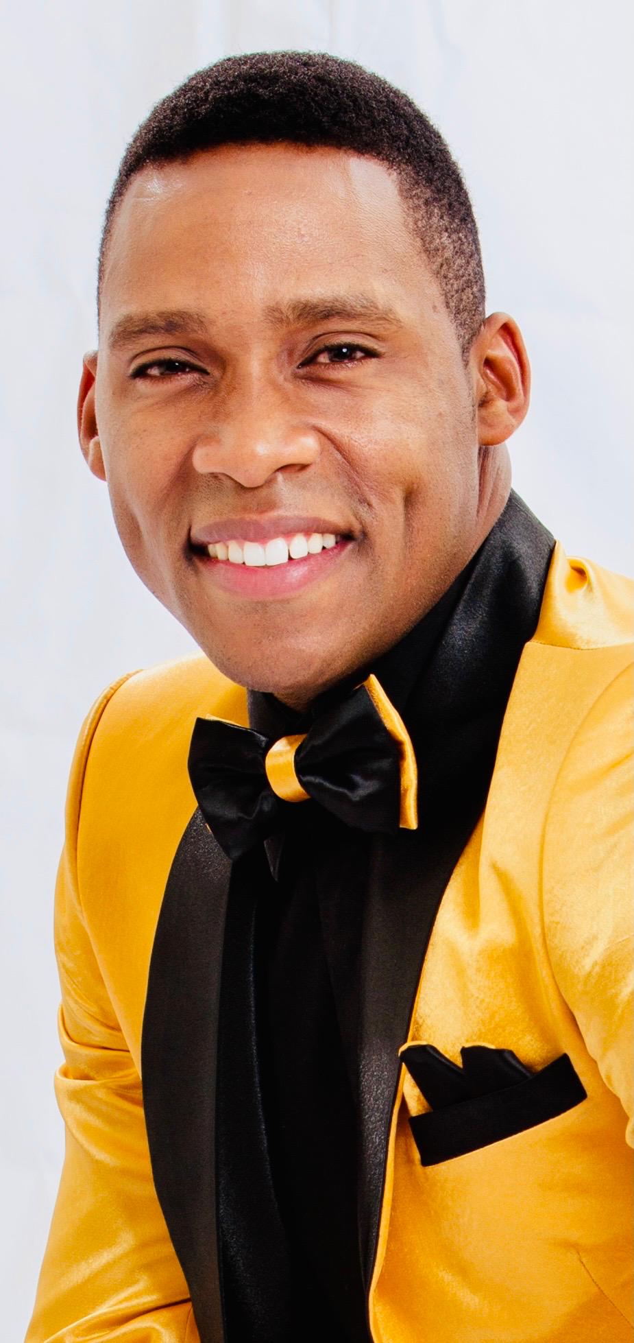 Man smiling, wearing a yellow jacket and black bow tie, against a light background.