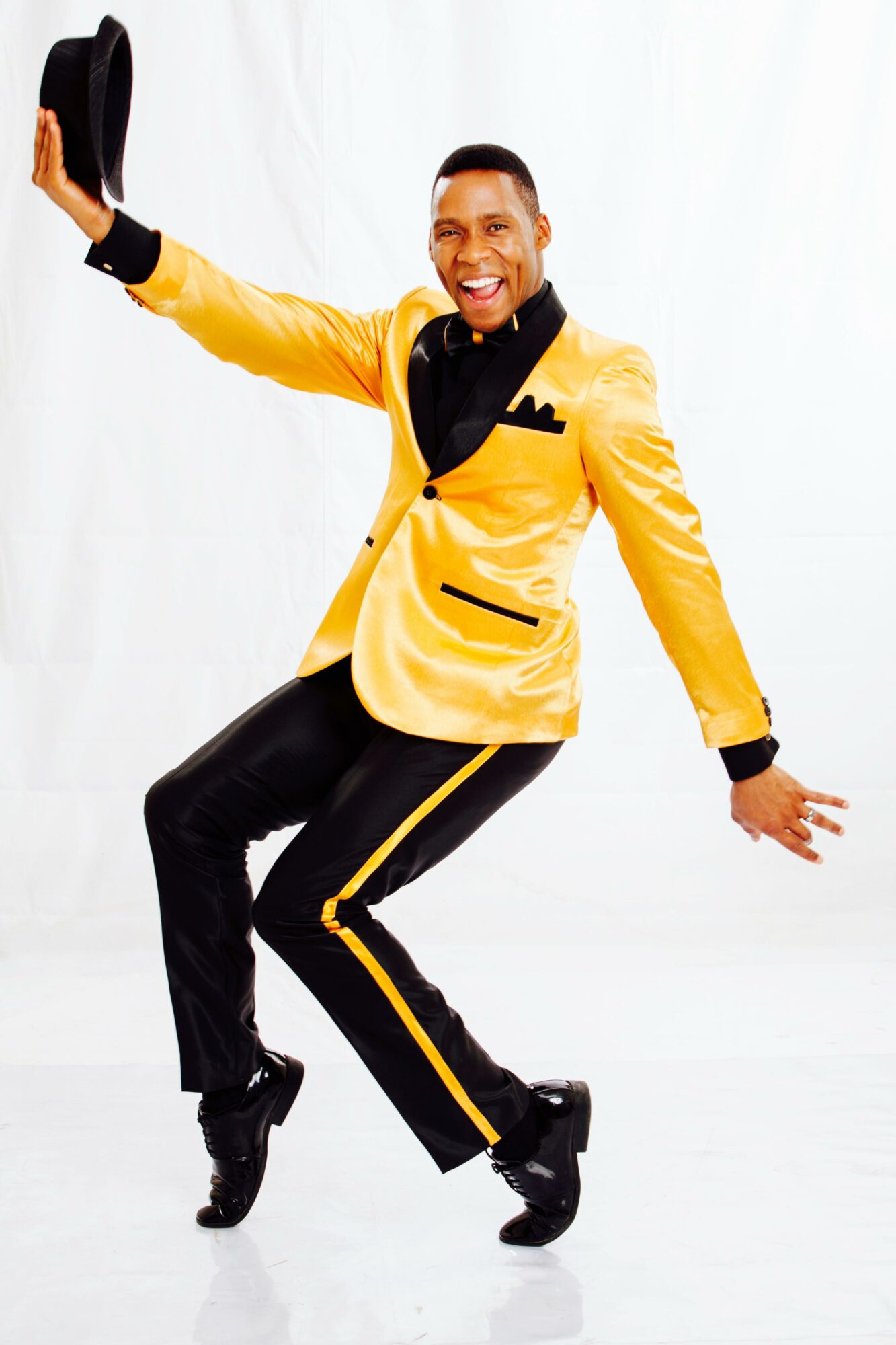 Person dancing, wearing a yellow jacket with black accents, black pants with yellow stripes, holding a hat, smiling, against a white background.
