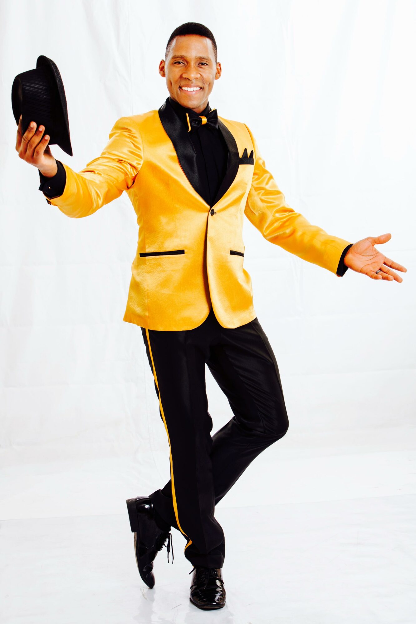 Man in yellow tuxedo with black bow tie holding a hat, smiling, with arms open, standing on white background.