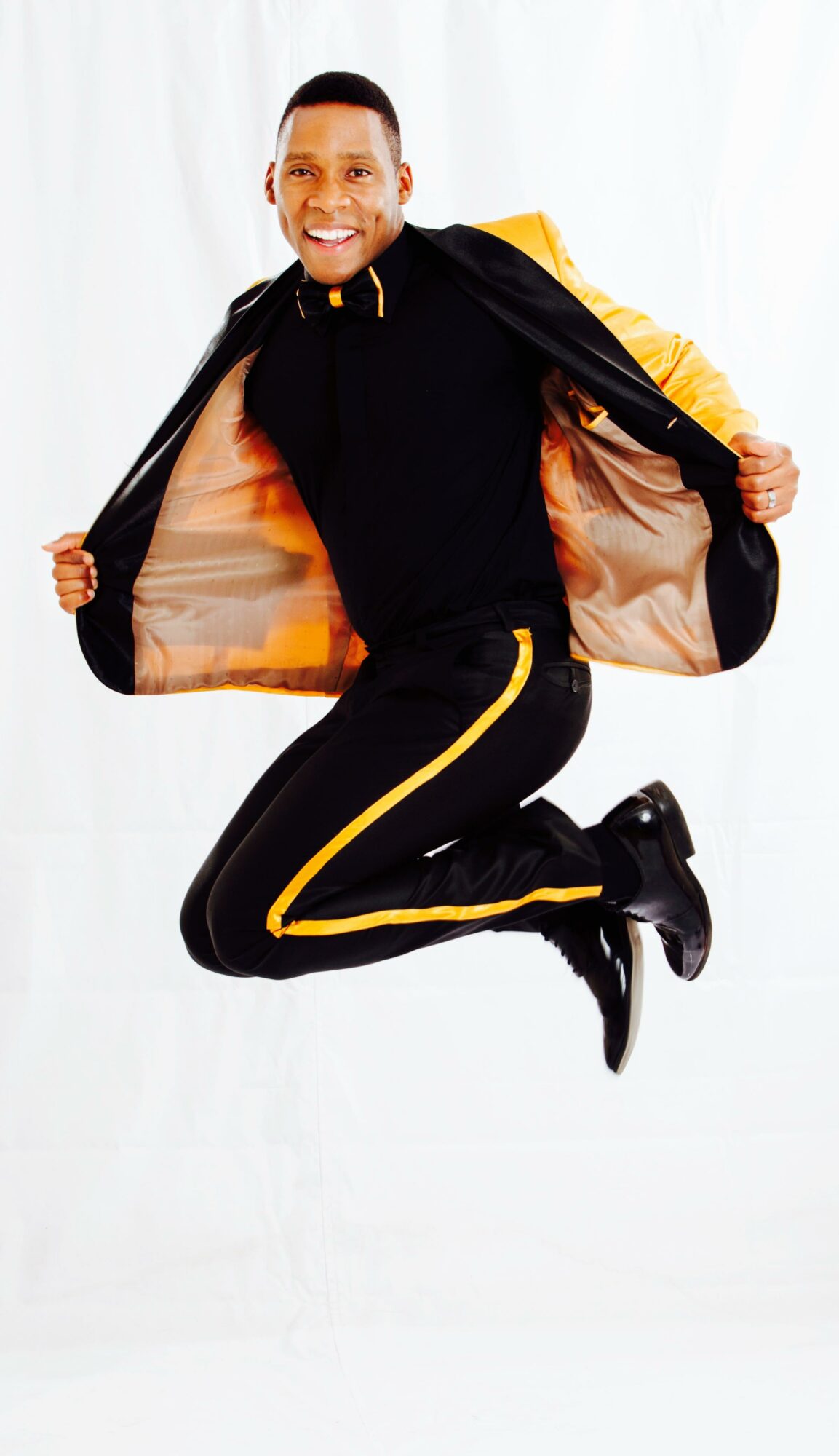 Person jumping with a black and yellow jacket, black pants with yellow stripe, and black shoes, smiling, white background.