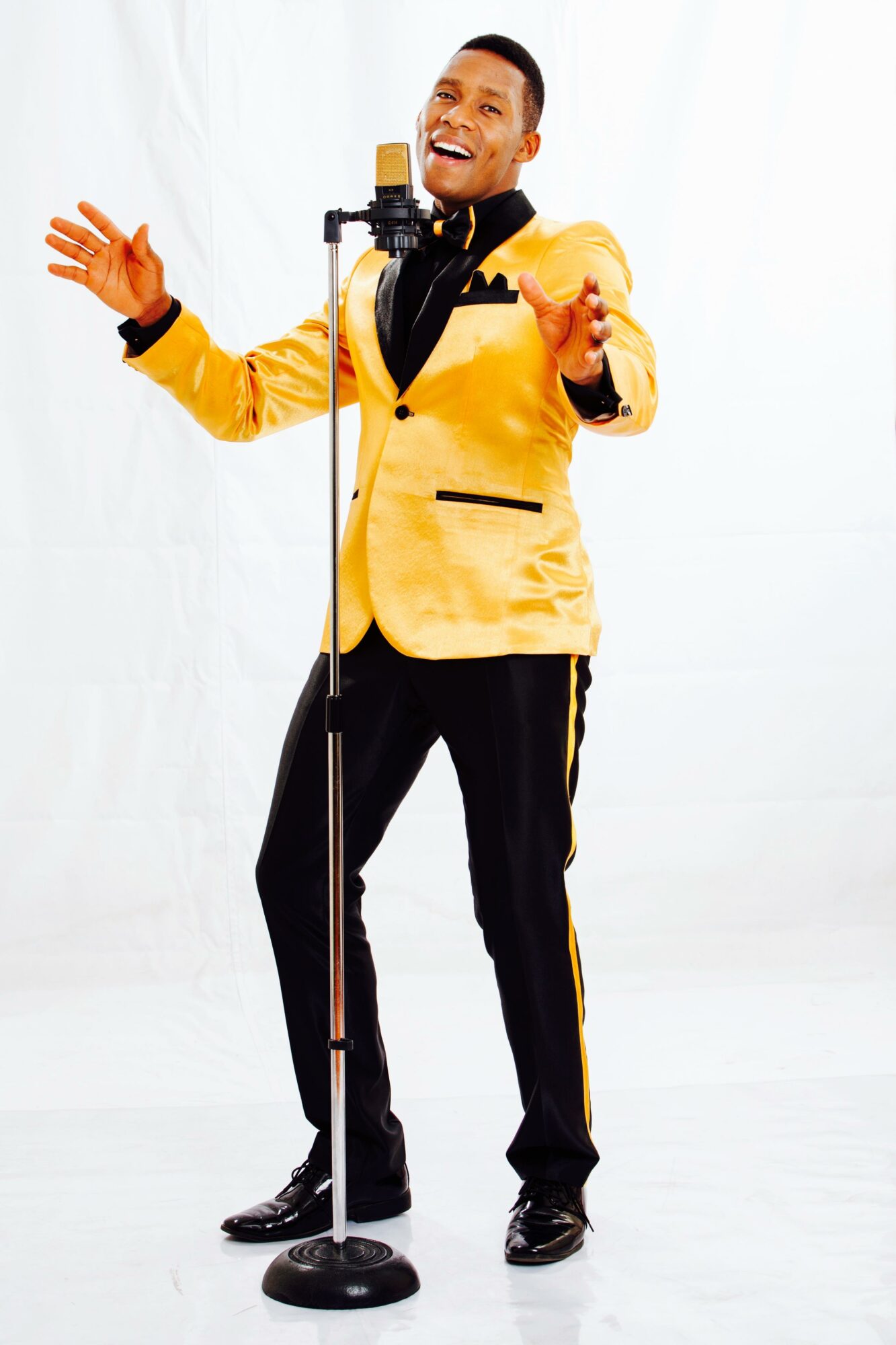 Man singing into a microphone with arms outstretched, wearing a yellow jacket and black pants, smiling.
