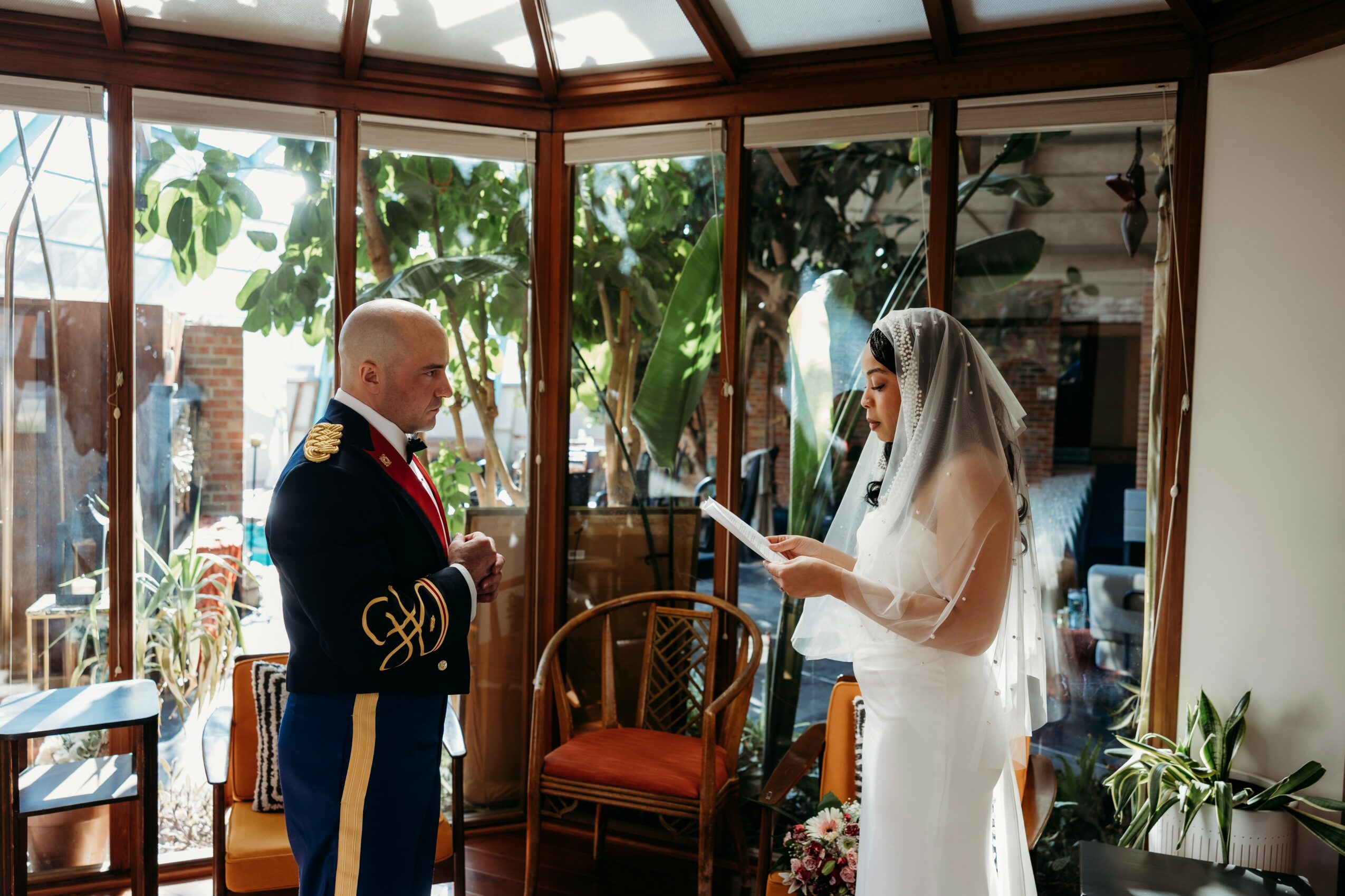 Bride and groom facing each other indoors with large windows and plants, bride reading vows, groom in uniform, warm natural light.