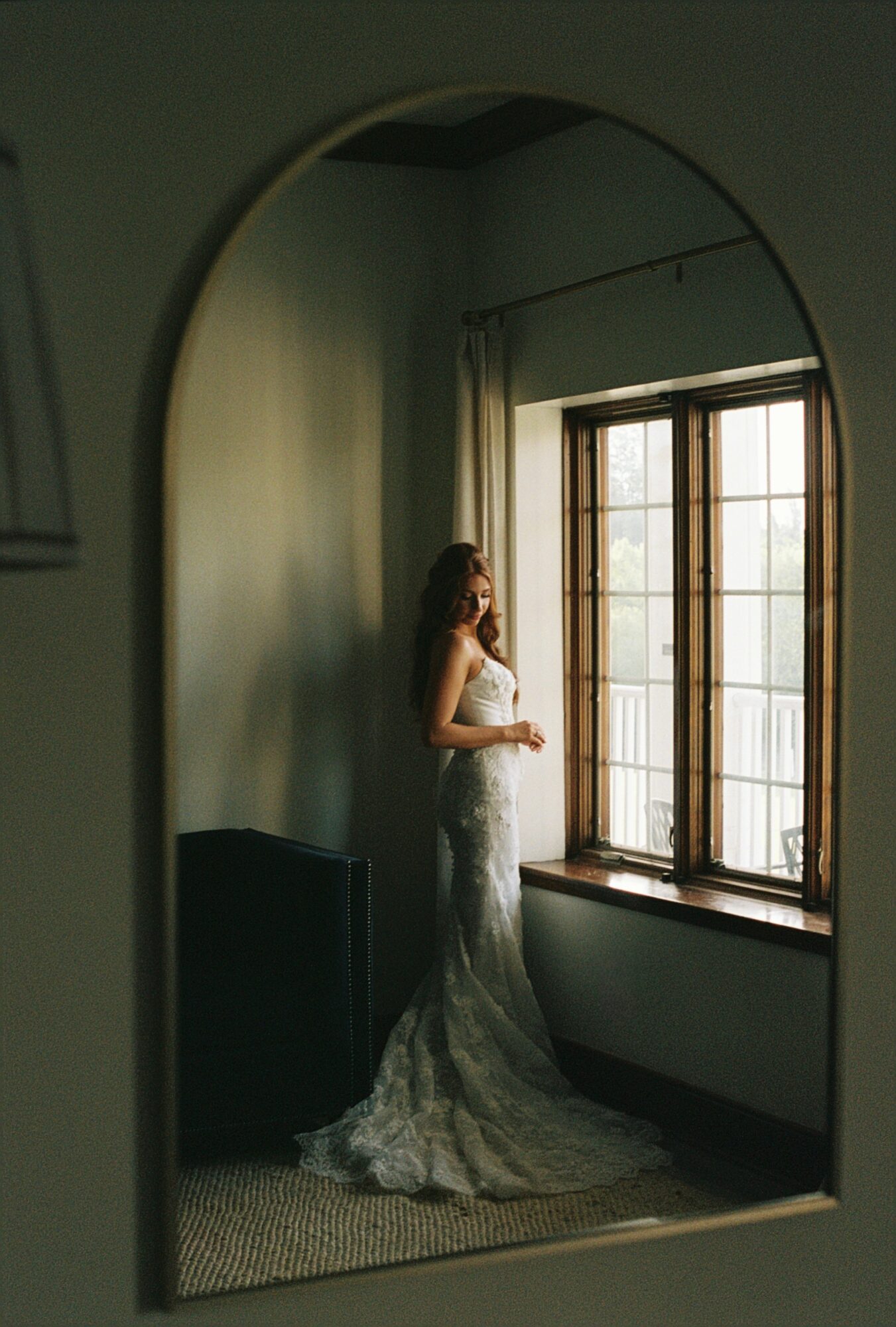 Woman in a wedding dress standing by a window in a room, reflected in an arched mirror.
