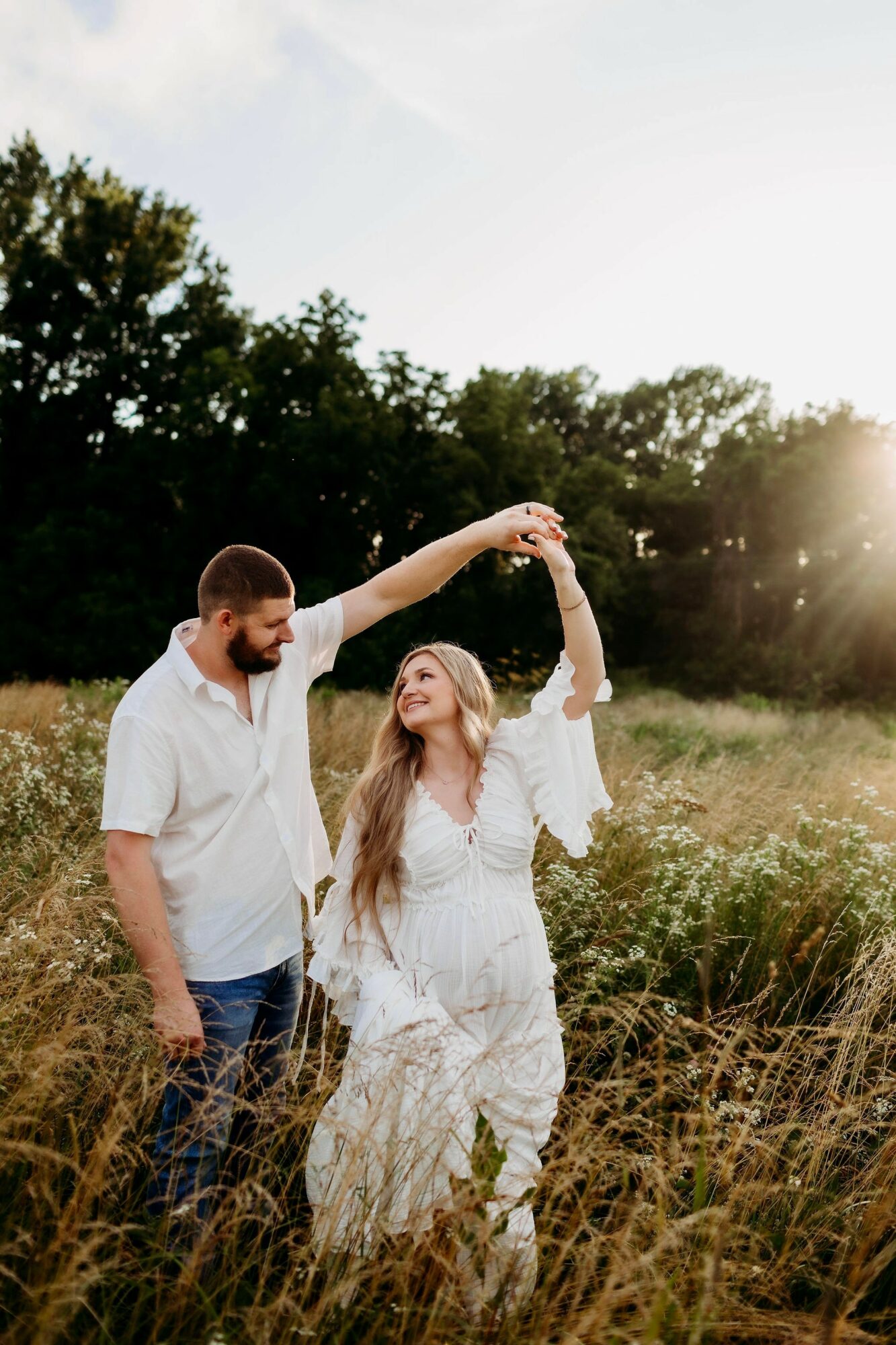 A man and woman dance in a field with trees in the background, sunlight shining behind them, woman wearing a white dress.