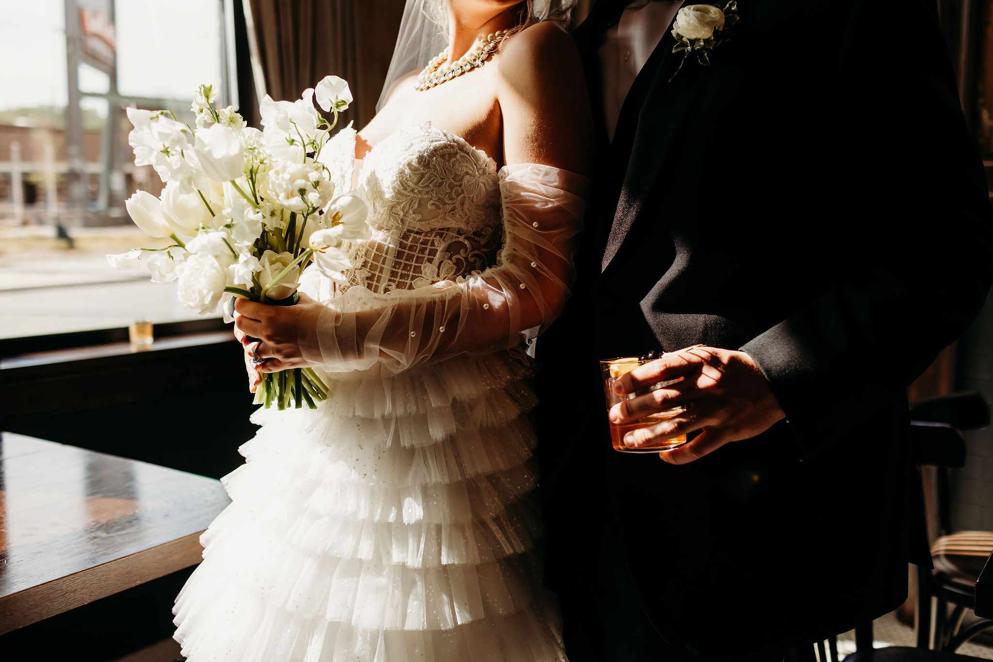 Bride holding a bouquet of white flowers, wearing a layered wedding dress, standing next to a person in a dark suit holding a glass.