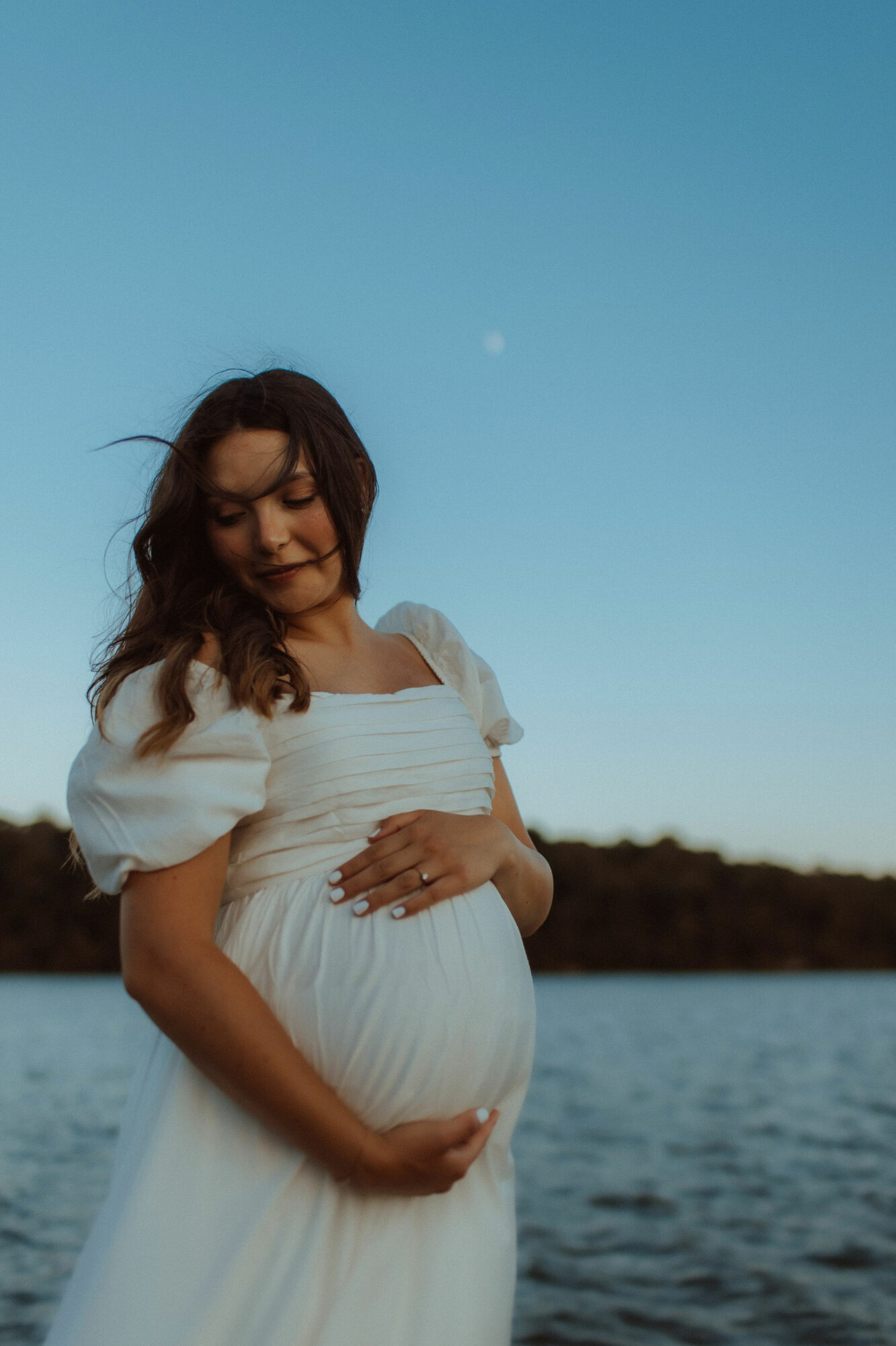 Pregnant woman in white dress standing outdoors near water, with a clear sky and moon in the background.