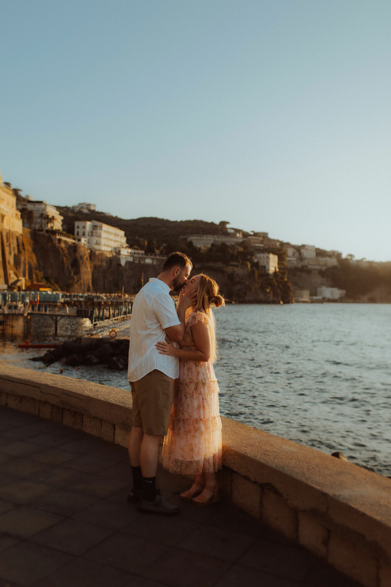 A couple stands close together by a waterfront, with buildings on cliffs in the background, during daytime.
