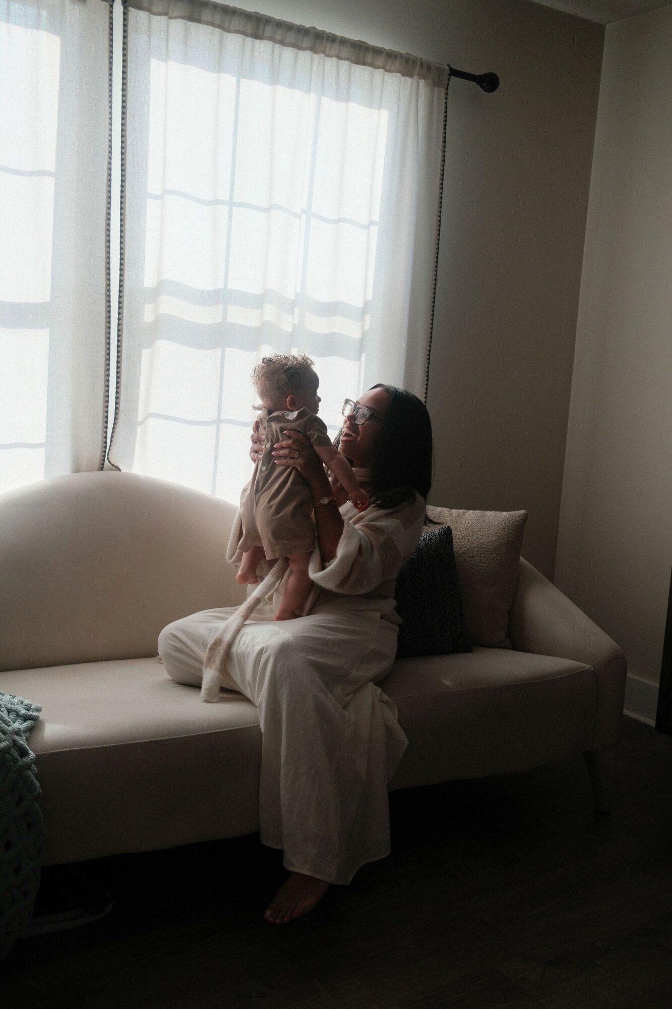 Person holding a child on a sofa near a window with curtains, natural light coming in, dark floor, light-colored walls.