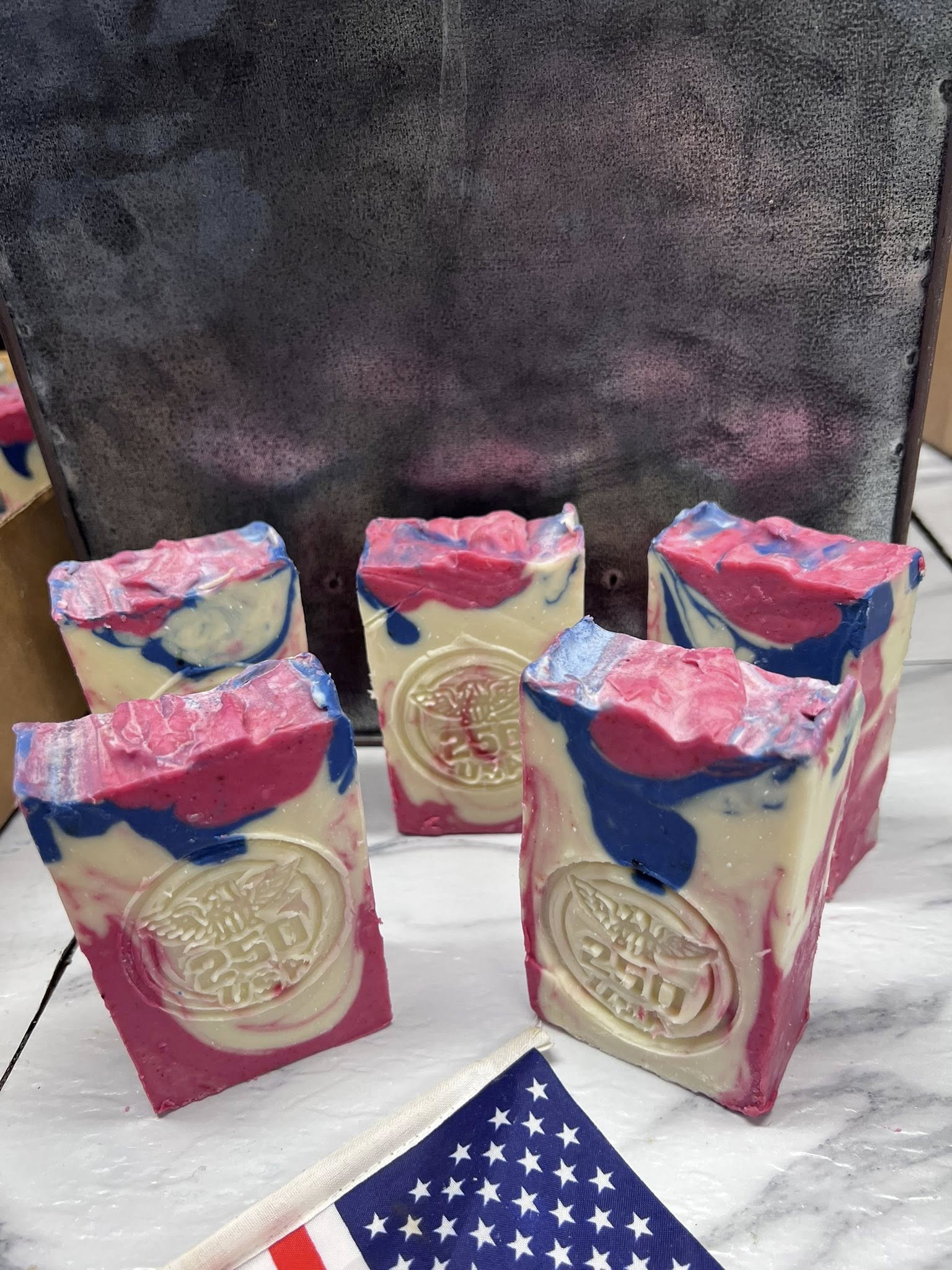 Six soap bars with colorful swirls and embossed designs, arranged on a marble surface with a dark background.