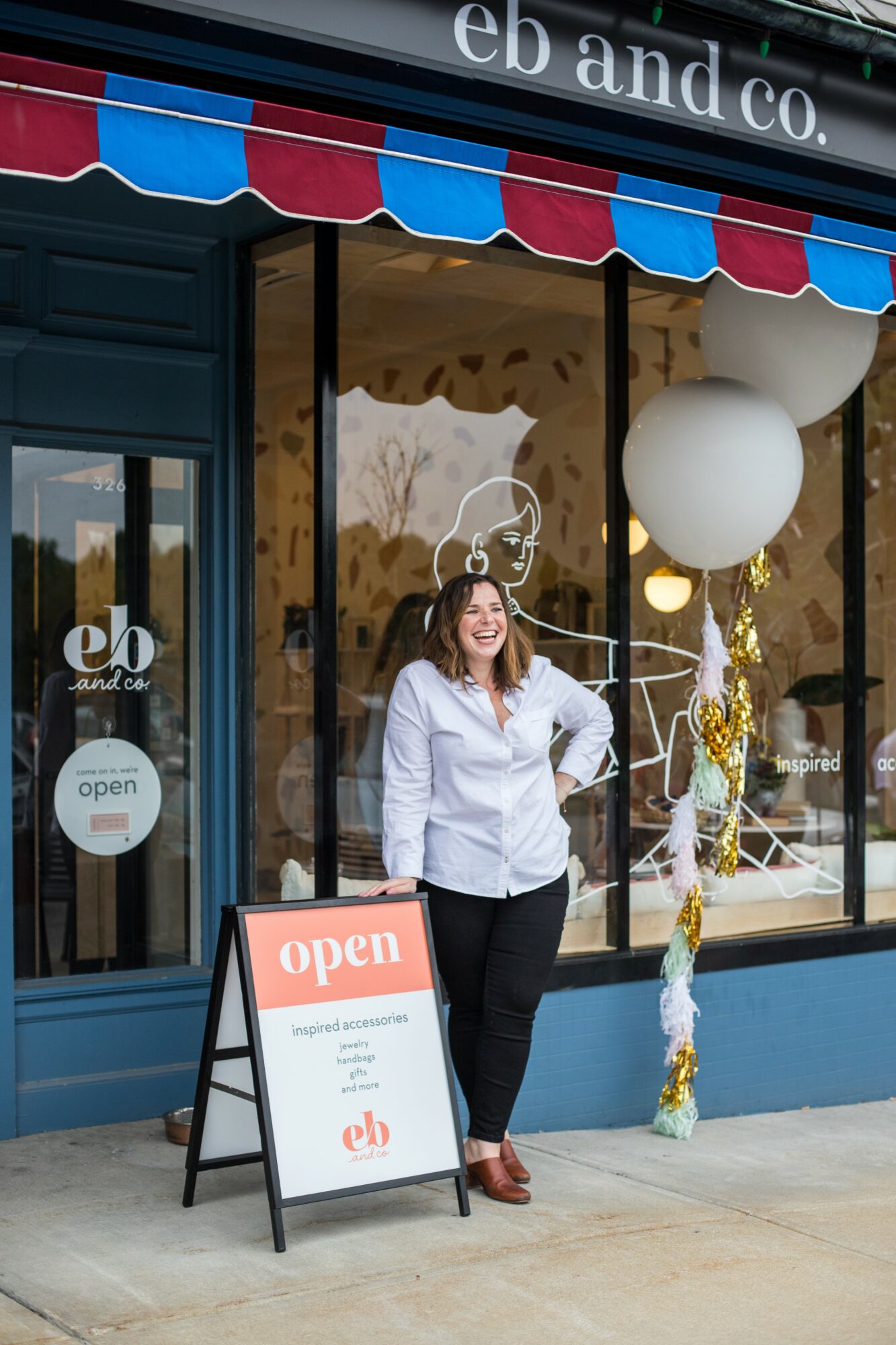 Woman standing outside storefront with an open sign, decorated with balloons and a festive display, smiling at camera.