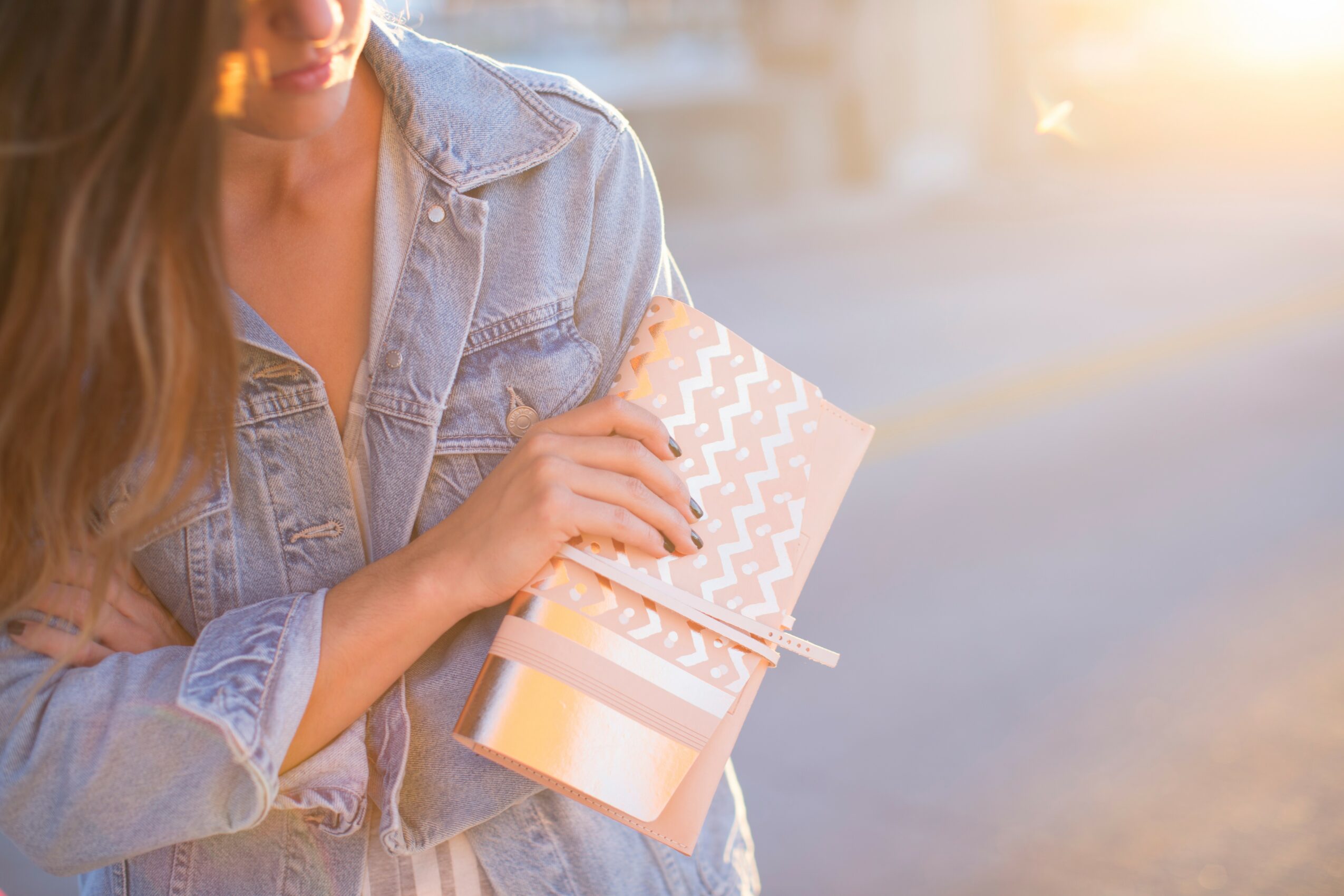 Person holding a small gift box with a patterned lid, wearing a denim jacket, outdoors during sunset.
