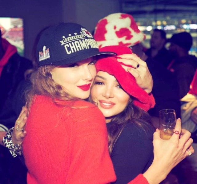 Two women hugging, one wearing a black cap with 'Champions' and a red jacket, the other in a red hat and black top, holding a drink.
