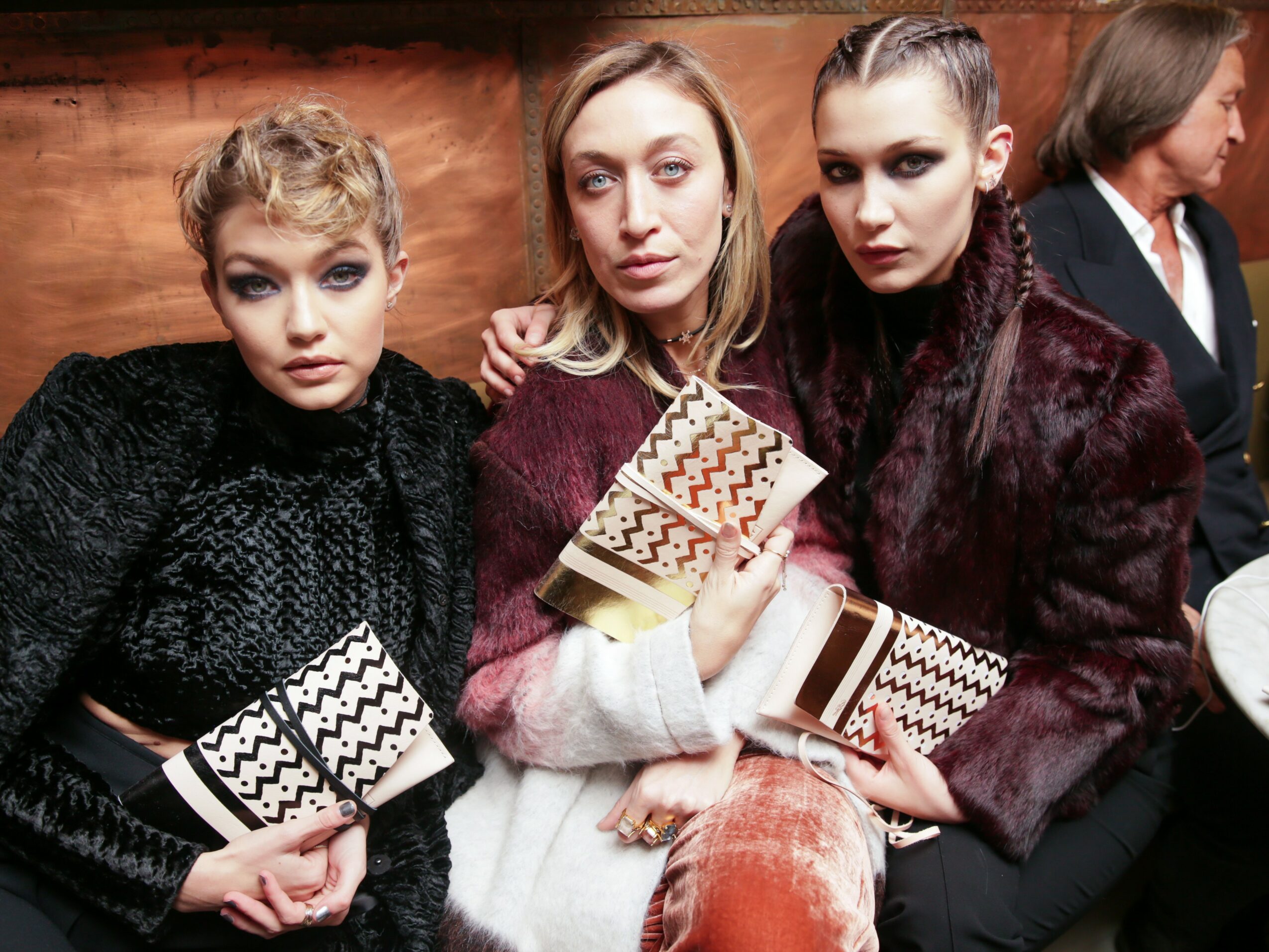 Three women sitting together, holding patterned clutch bags, with a man in the background, in an indoor setting.