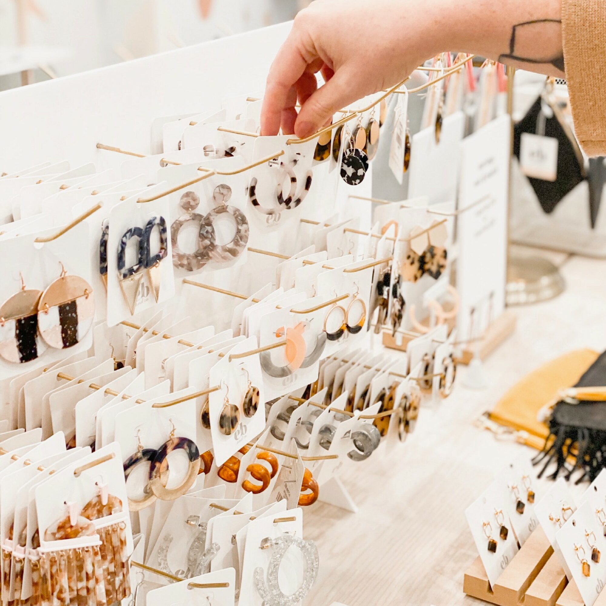 Hand reaching for earrings displayed on a jewelry stand with multiple rows of earrings on cards. Bright, well-lit setting.