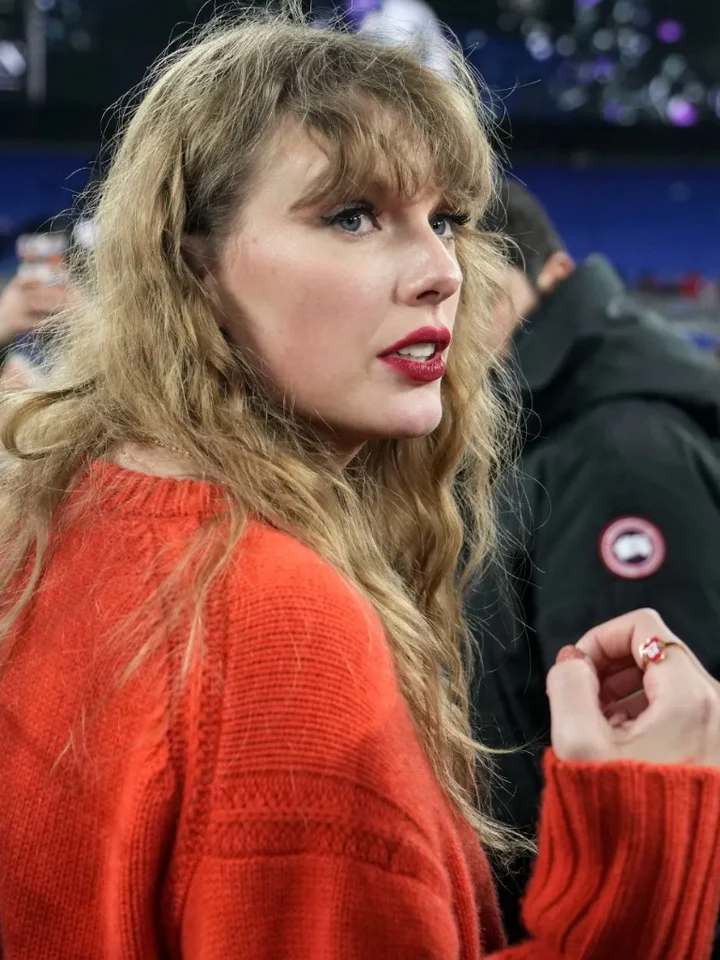 Woman with wavy blonde hair wearing a red sweater, looking to the side, in a crowded indoor setting.